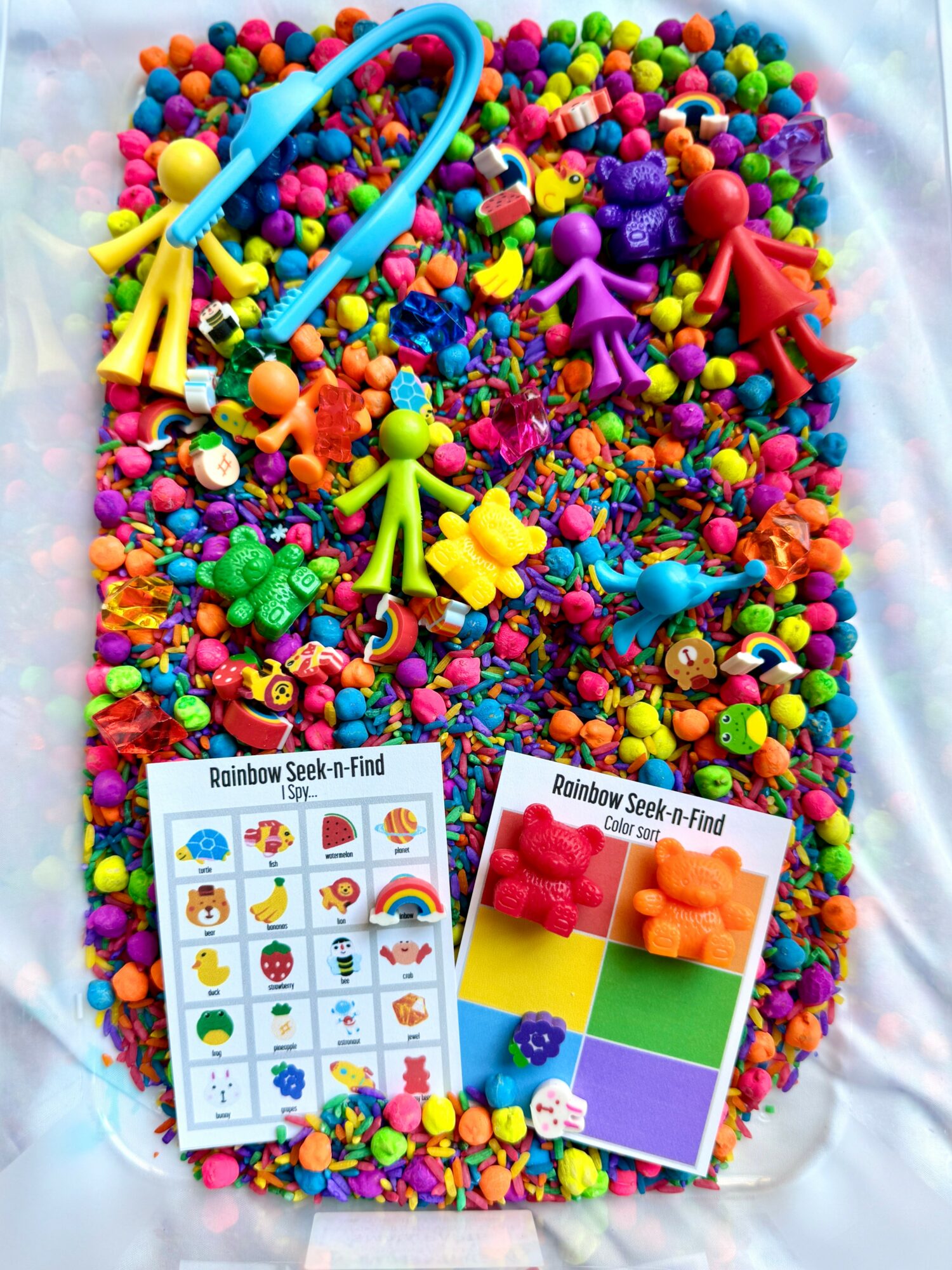 Colorful beads, toys, and two Rainbow Seek-n-Find activity cards on a white surface.