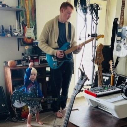 Young person playing electric guitar in a music studio with various instruments and equipment around.