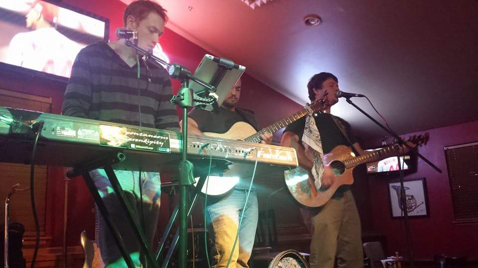 Two musicians perform on stage, one playing keyboard and the other singing and playing guitar, in a dimly lit venue.