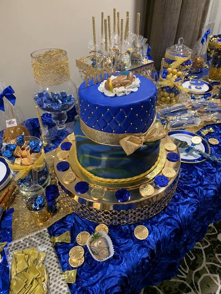 Two-tiered blue and gold cake with gold bows, surrounded by blue and gold decorations on a table.