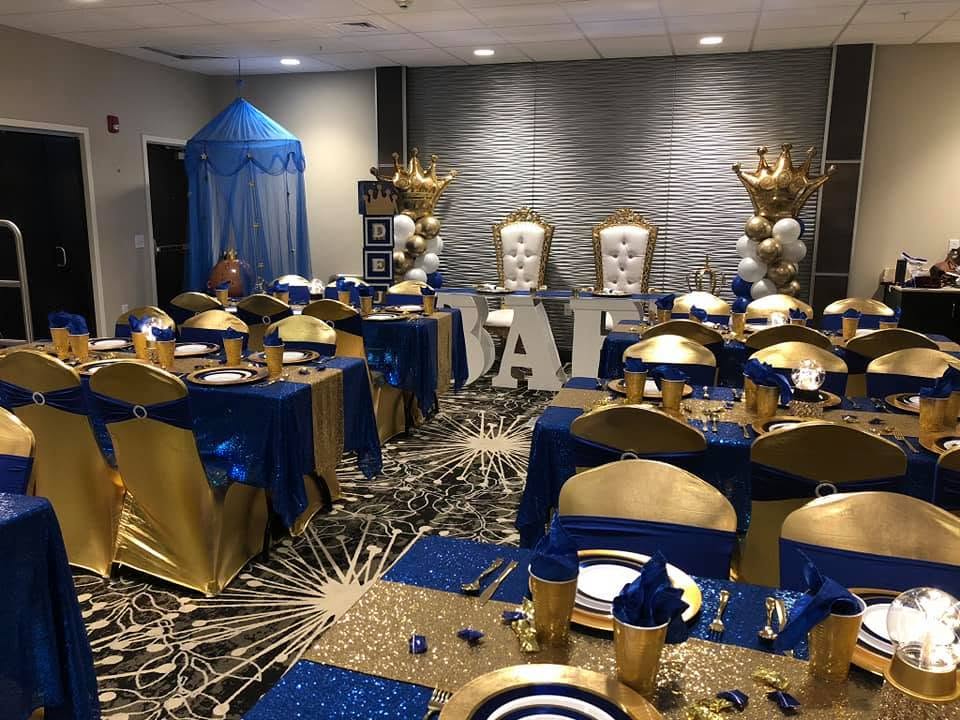 Party setup with decorated tables, chairs, and a stage with balloons and large crowns, in blue, gold, and white colors.