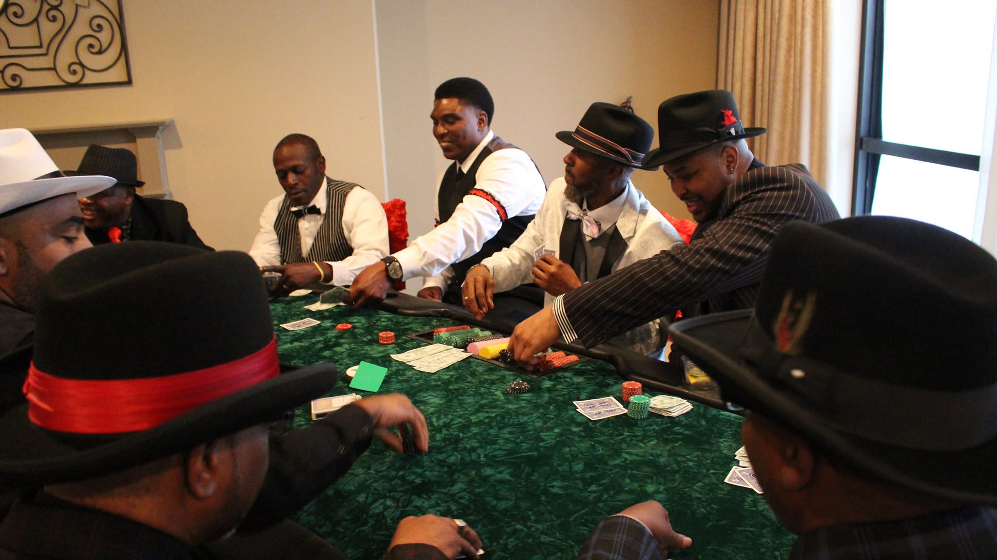 Group of men playing poker around a green table, some wearing hats and suits, in a room with a window.
