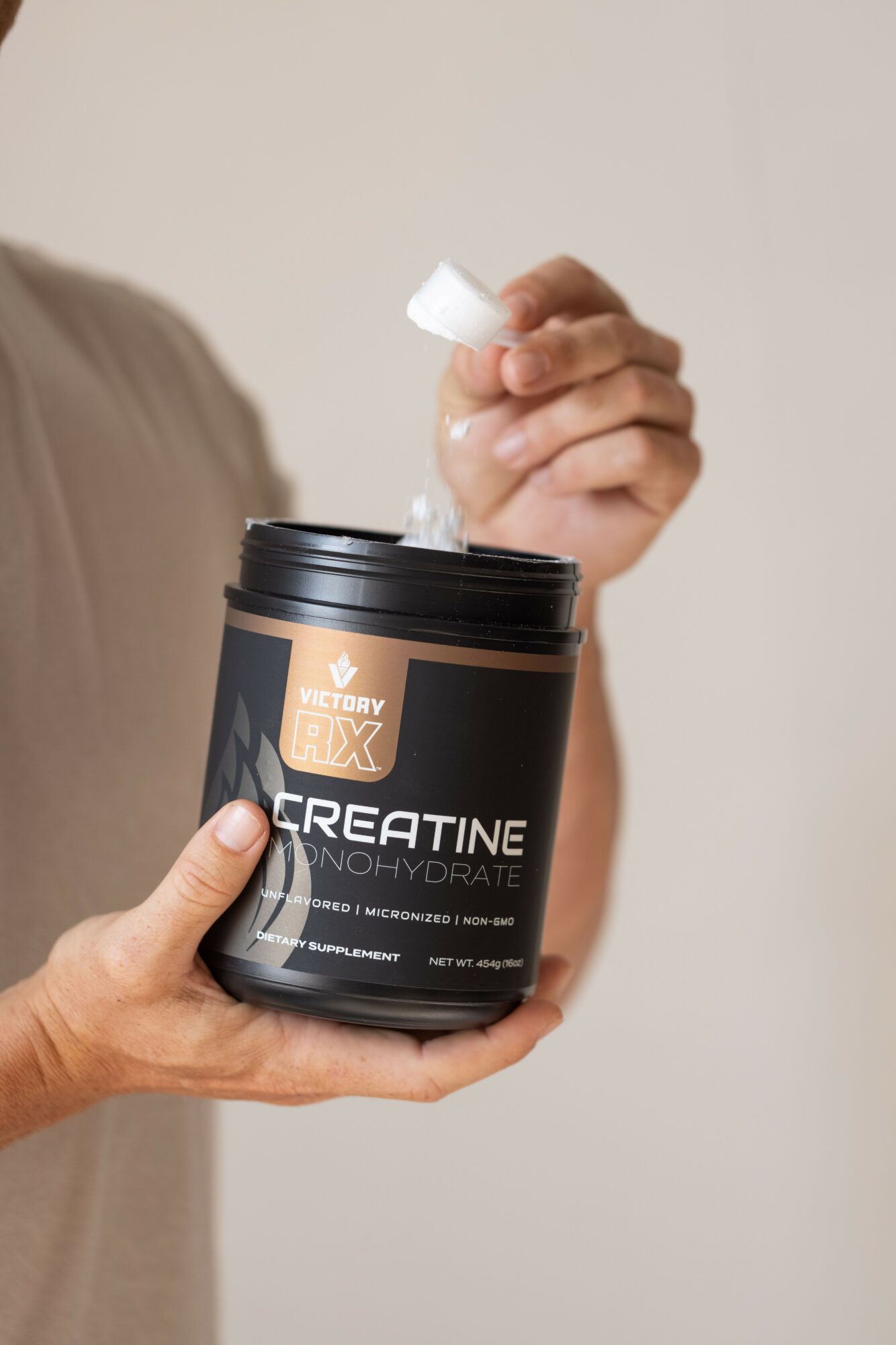 Person holding a black container of creatine supplement, pouring powder into hand, neutral background.