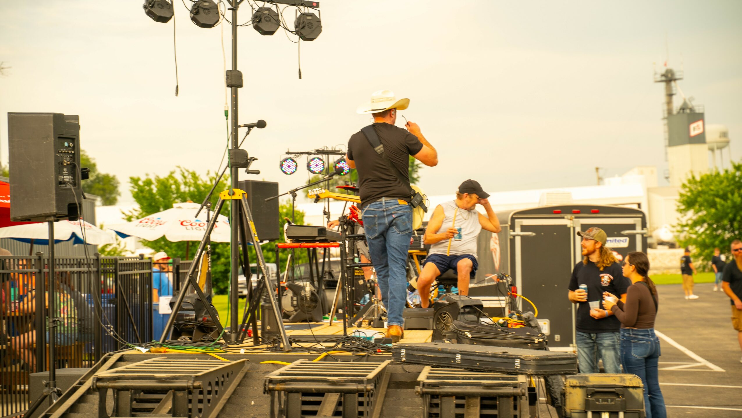 People on a stage with sound equipment and a person wearing a hat, outdoors during daytime.