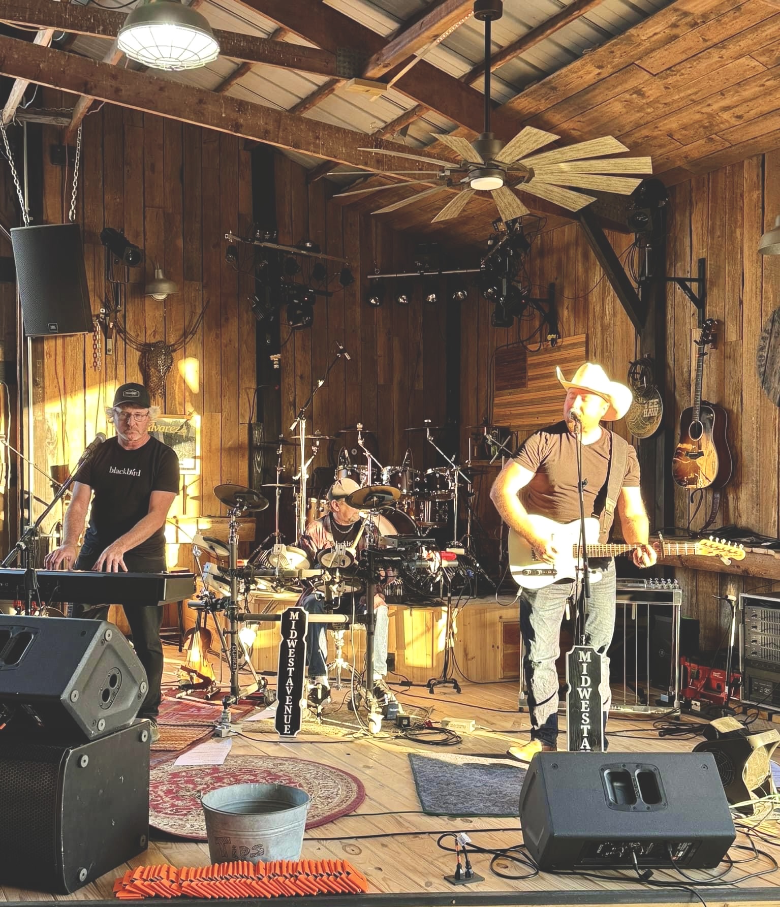 Three musicians perform on a wooden stage with instruments and equipment, wooden walls, ceiling fan, and stage lights, in a rustic setting.