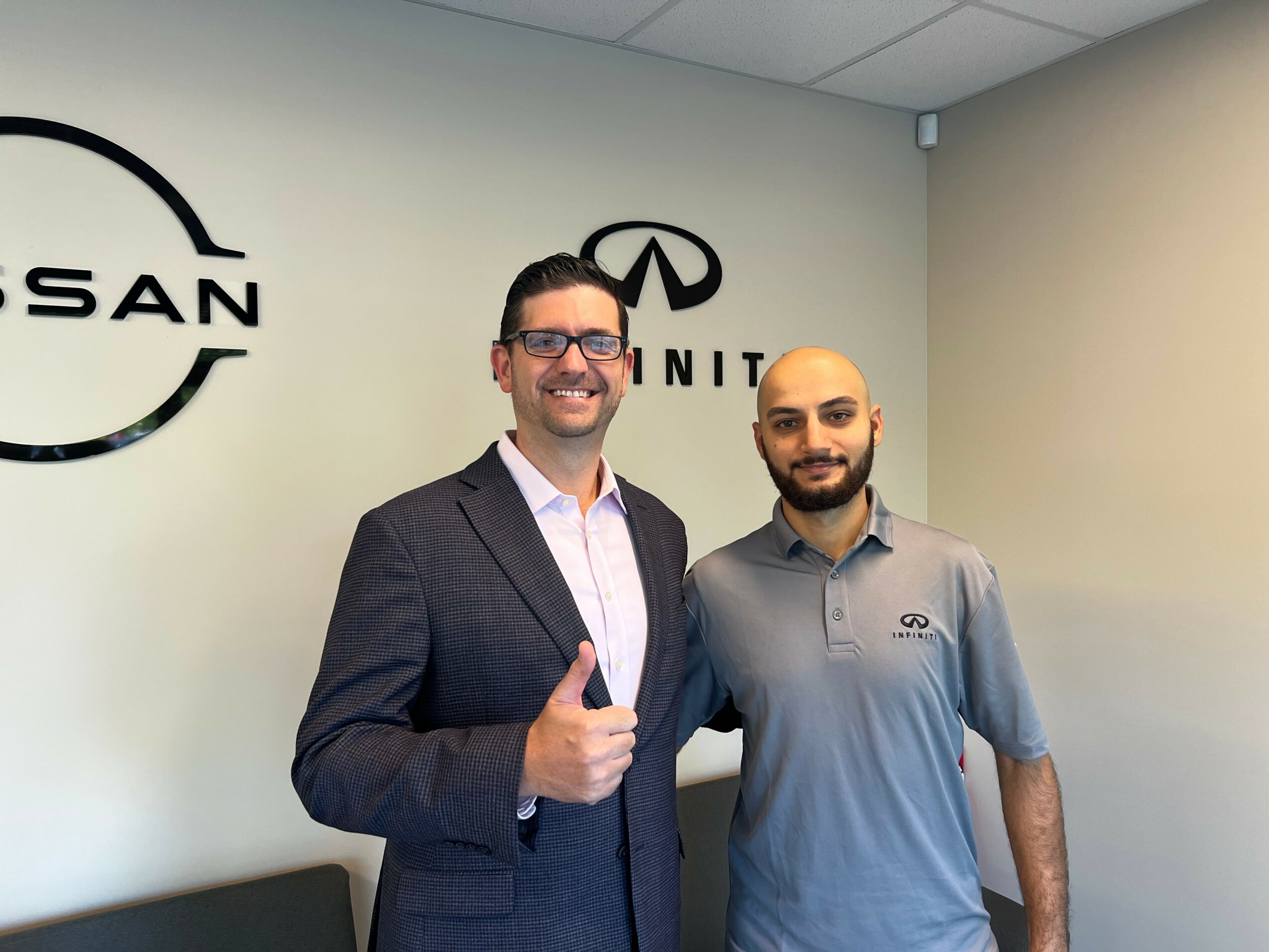 Two men standing indoors, one in a suit giving a thumbs-up, the other in a polo shirt, with Nissan and Infiniti logos on the wall behind them.