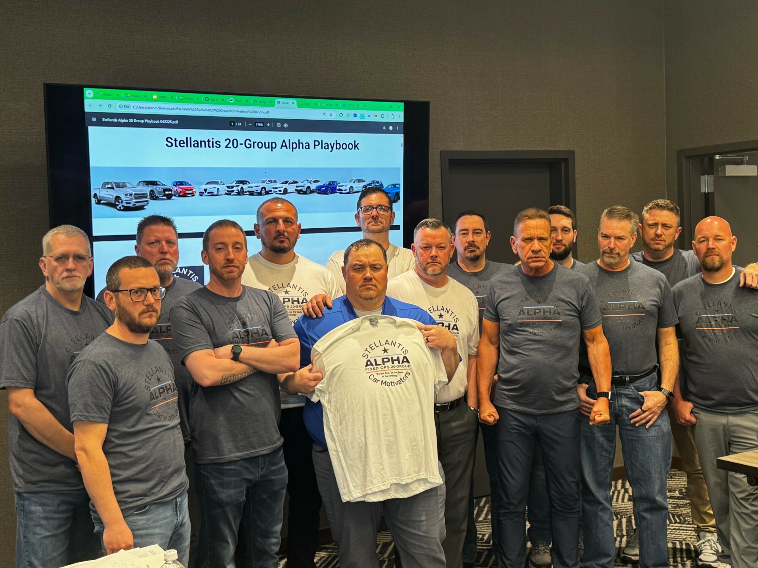 Group of fifteen men standing indoors, some wearing matching gray T-shirts, with a screen displaying text behind them.