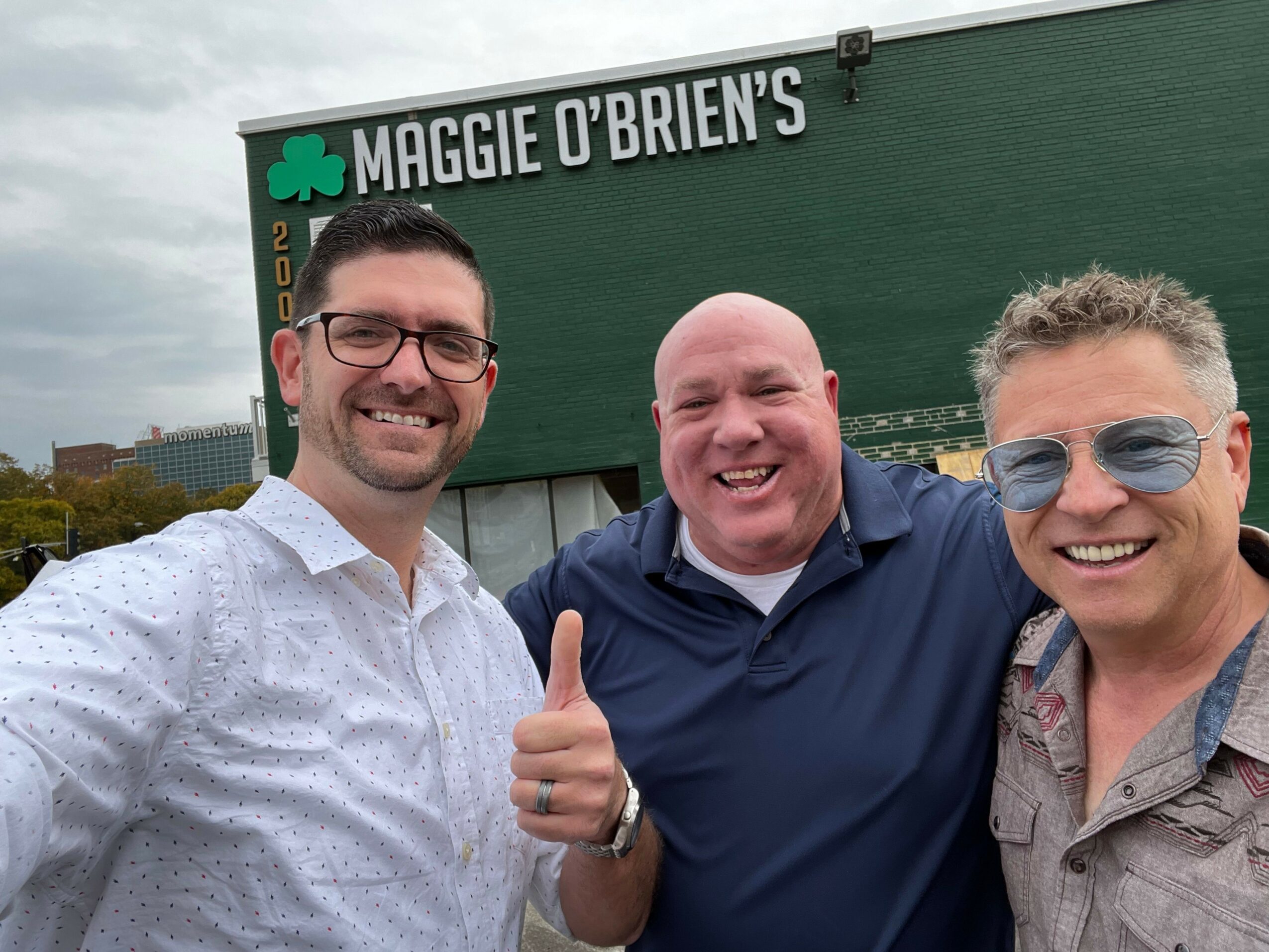 Three people smiling and giving thumbs up outdoors, standing in front of a green sign with white text.