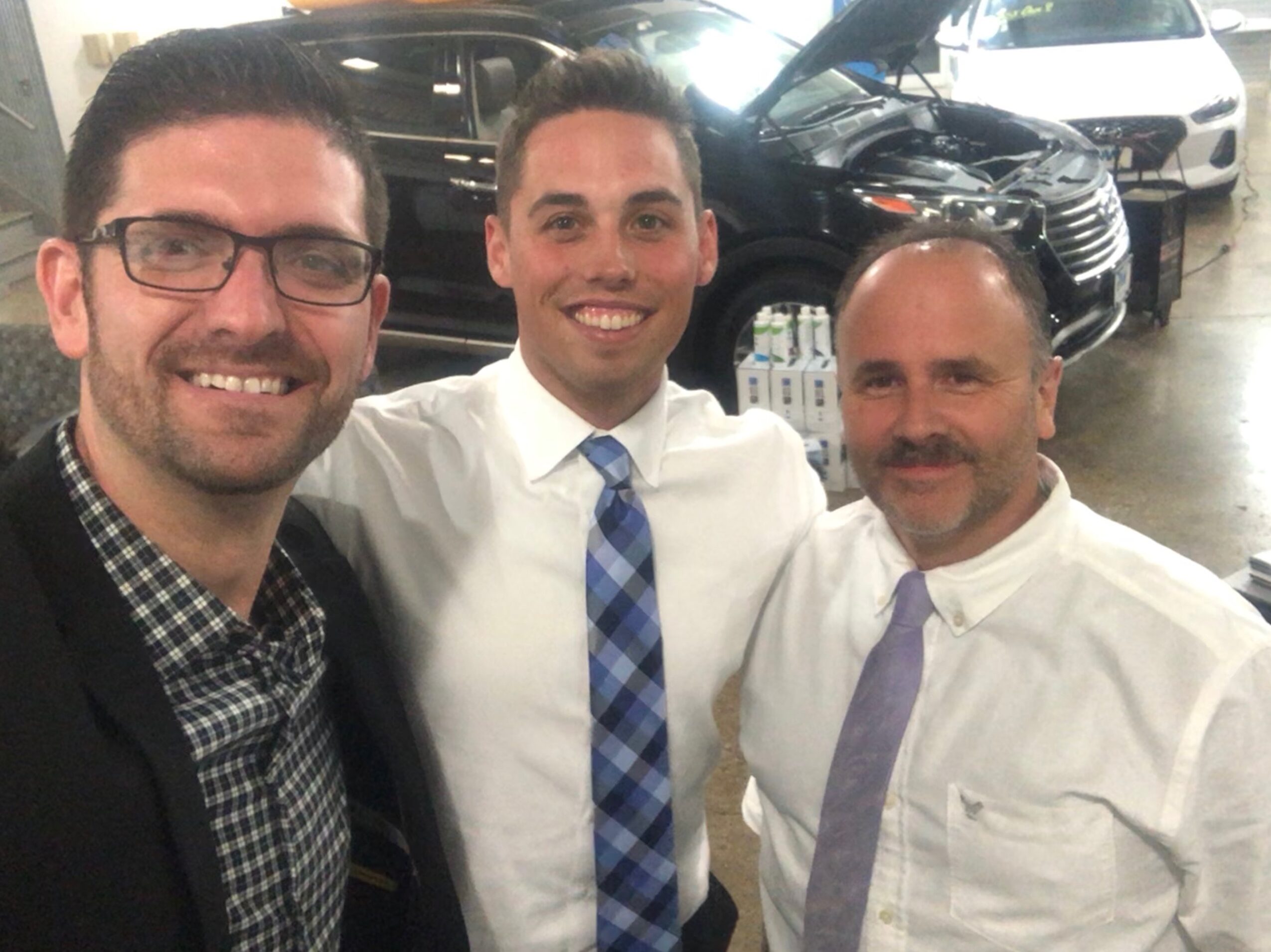 Three men smiling in a parking garage, dressed in formal attire, with cars in the background.