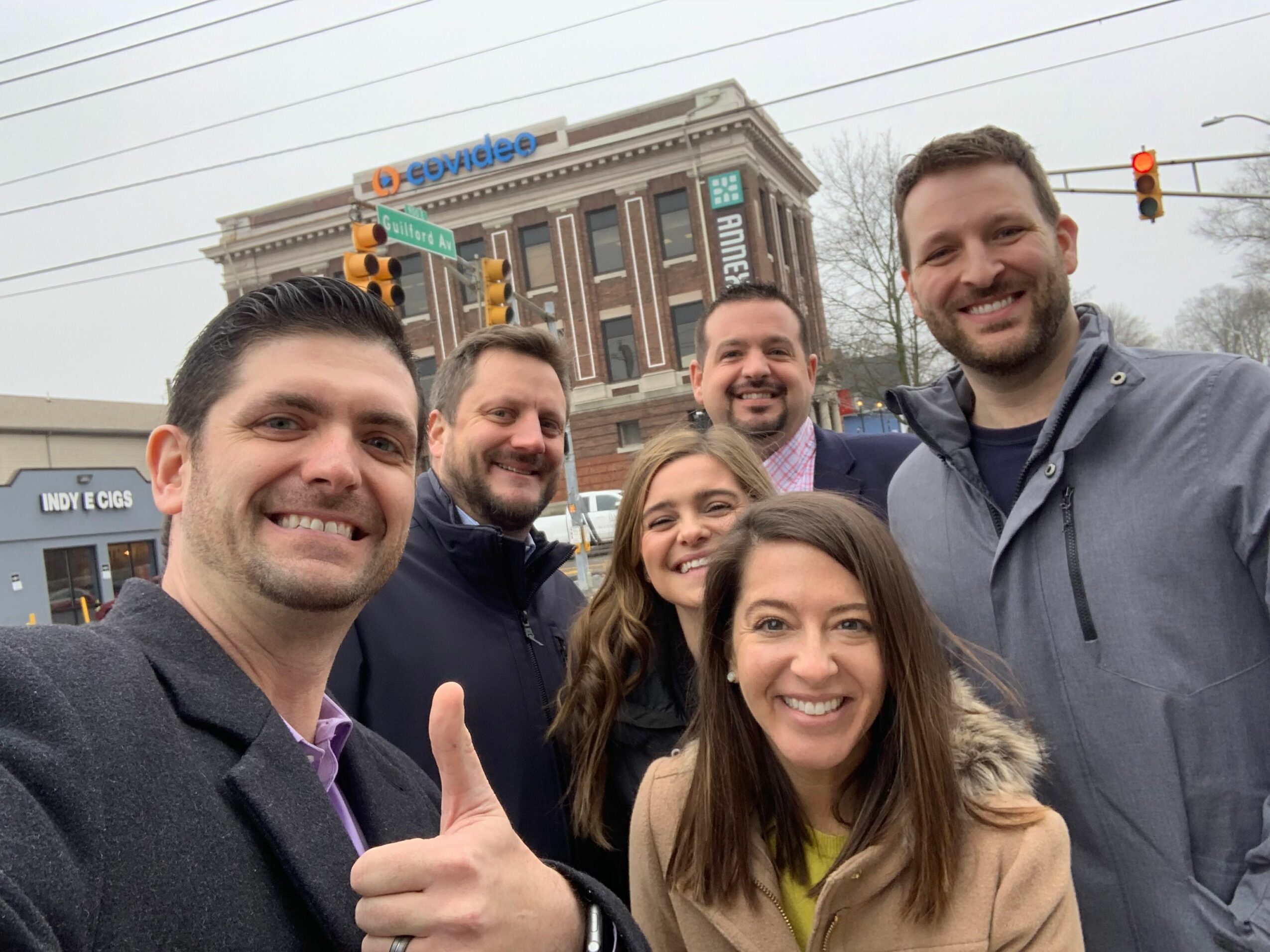 Group of seven people smiling and posing outdoors in front of a city street with buildings and traffic lights.