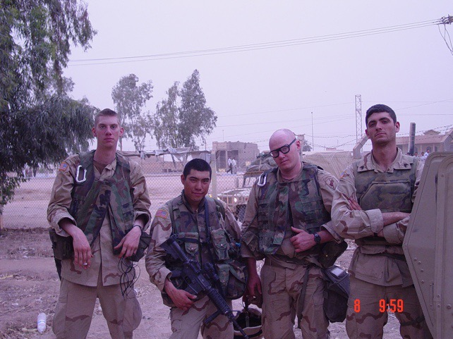 Four soldiers in uniform standing outdoors with trees and buildings in the background.