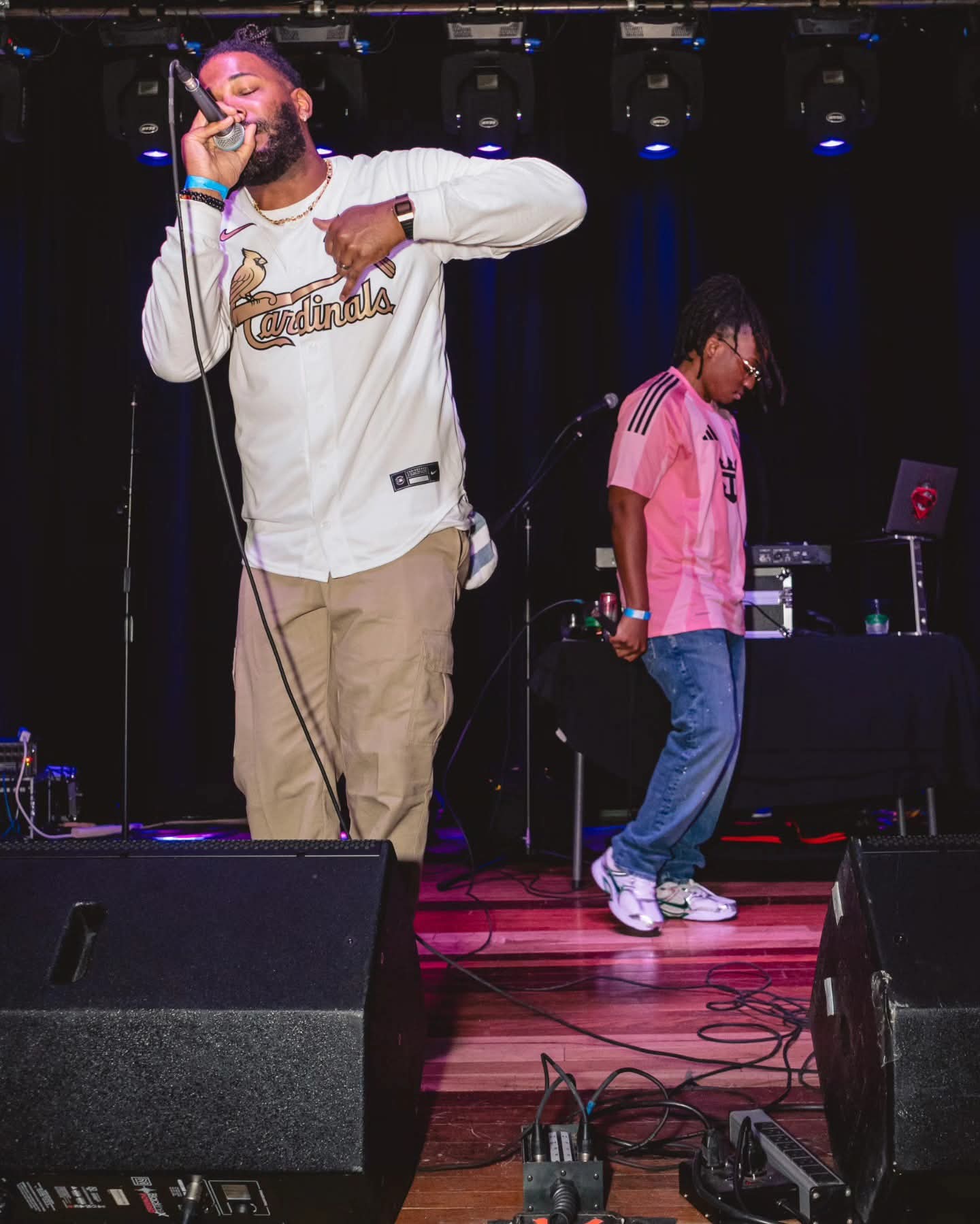 Two performers on stage, one singing into a microphone and the other standing near a DJ setup, with stage equipment visible.