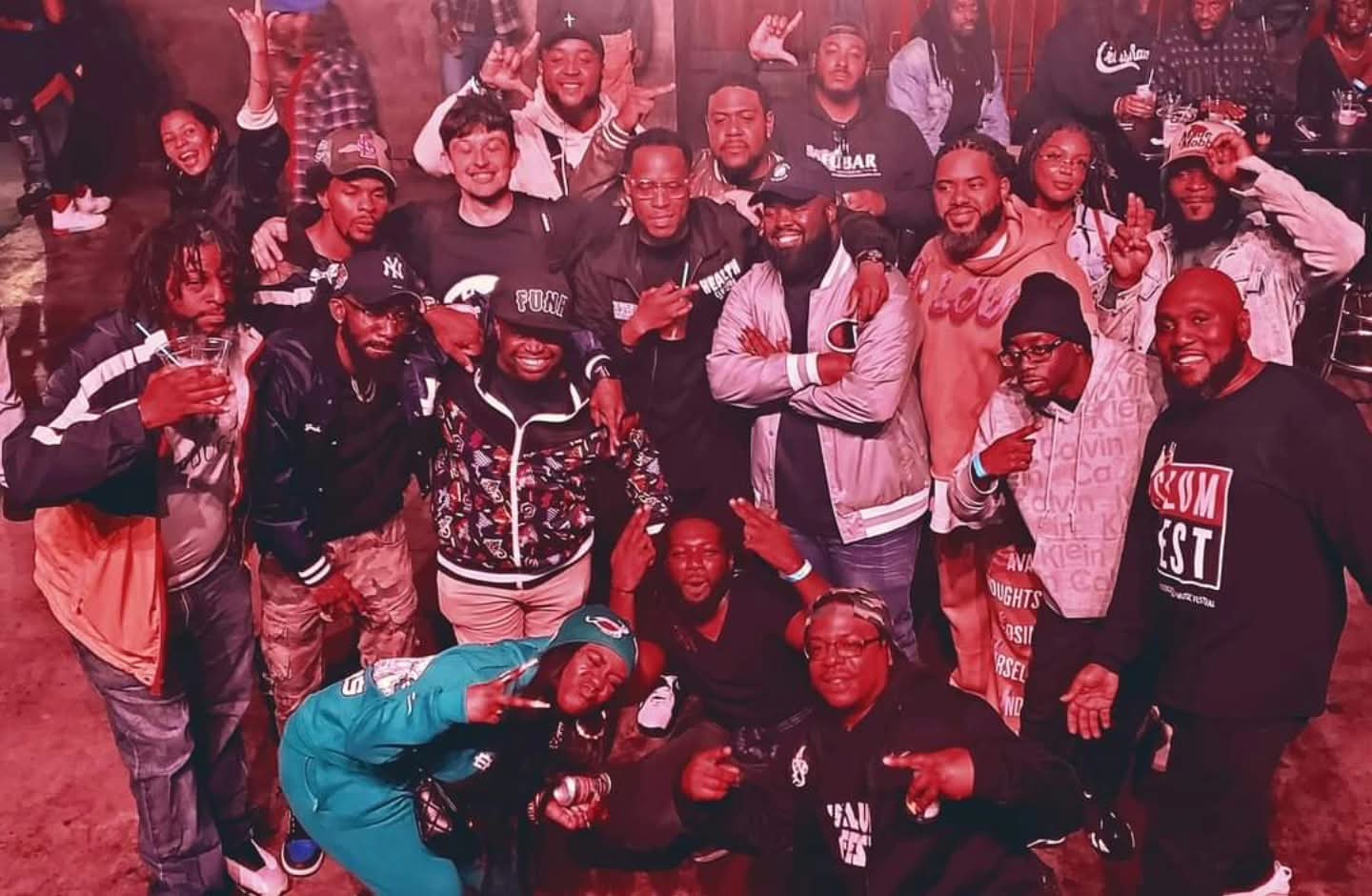 Group of people gathered indoors, some smiling and raising hands, with a dark background and casual clothing.