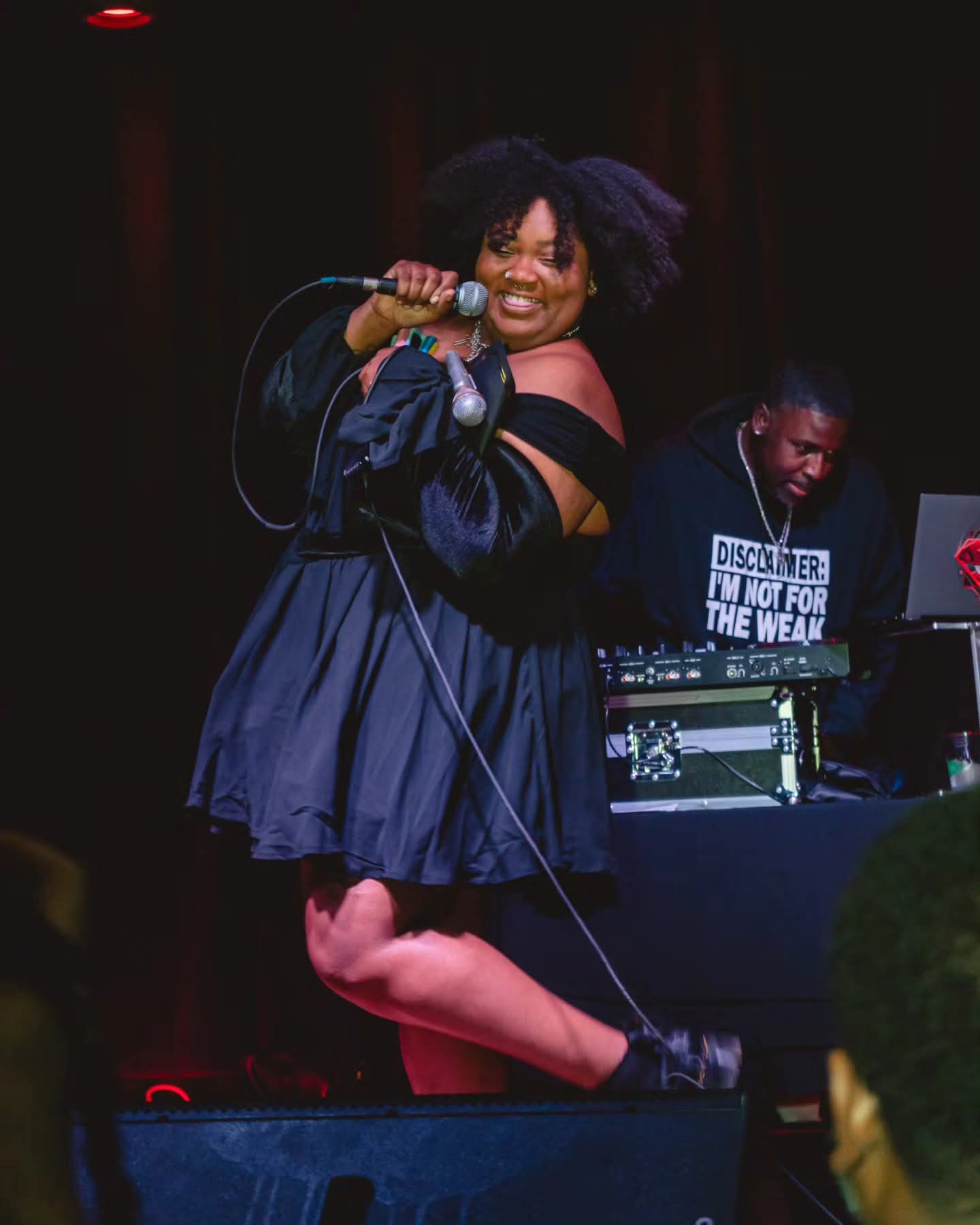 A woman singing into a microphone on stage, with a DJ in the background, smiling and wearing a dark dress.