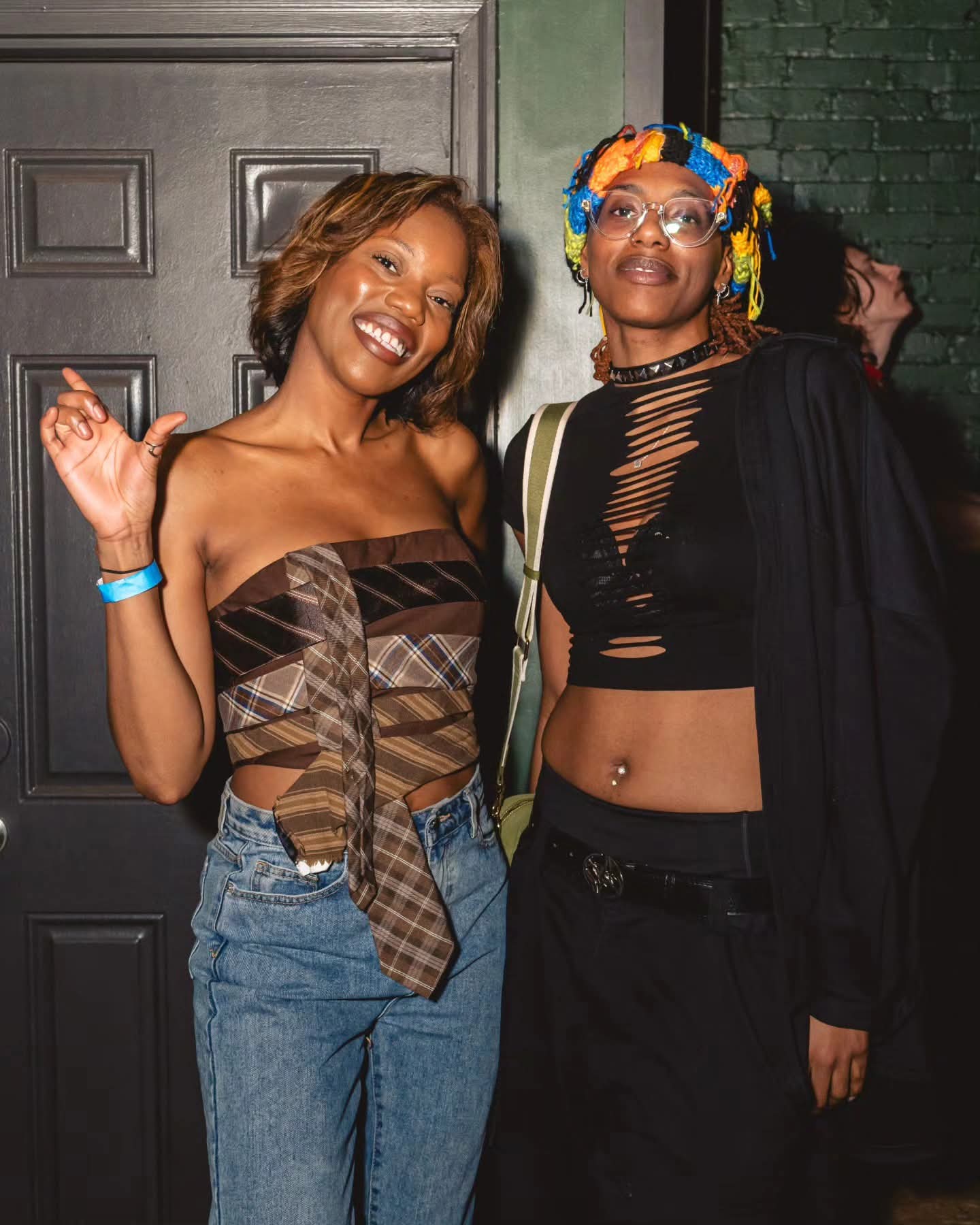 Two women smiling and posing together indoors, one with short hair and the other with colorful headwrap, both dressed casually.