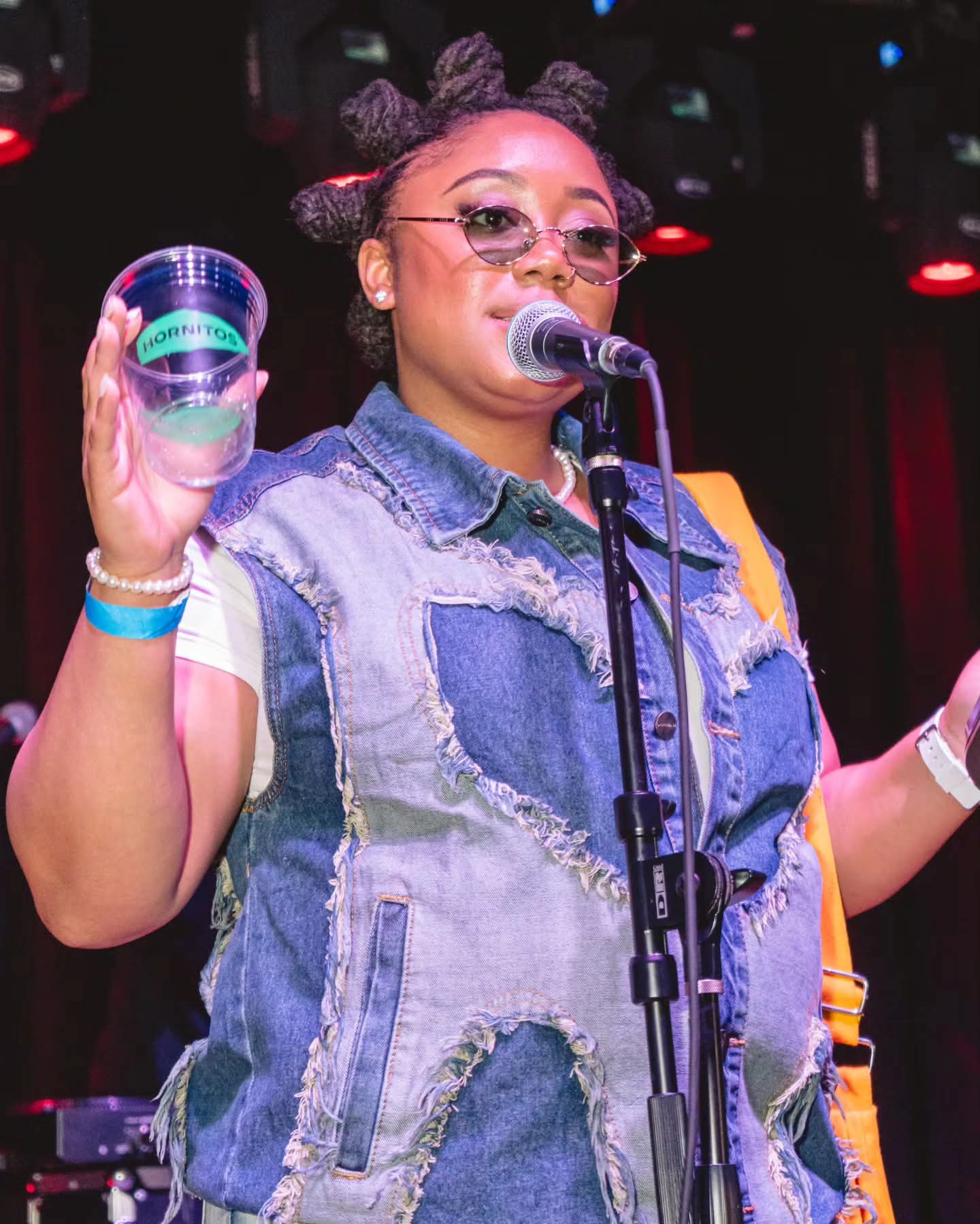 Person with styled hair, wearing sunglasses and a denim vest, holding a glass and speaking into a microphone on stage.