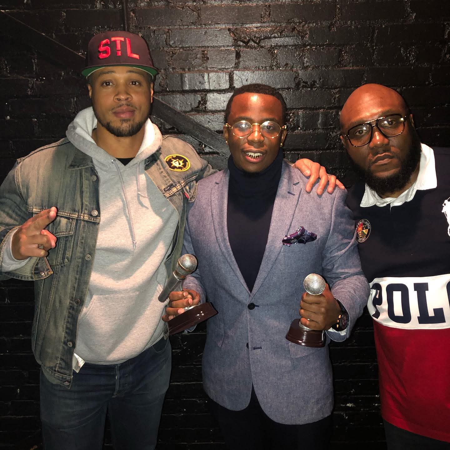 Three men standing against a black brick wall, two holding microphones, one wearing glasses and a suit, others casual clothing.