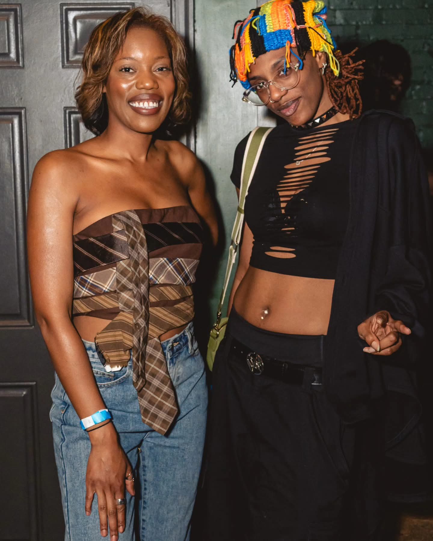 Two women smiling, one wearing a strapless top and jeans, the other in a black crop top and colorful headwrap.