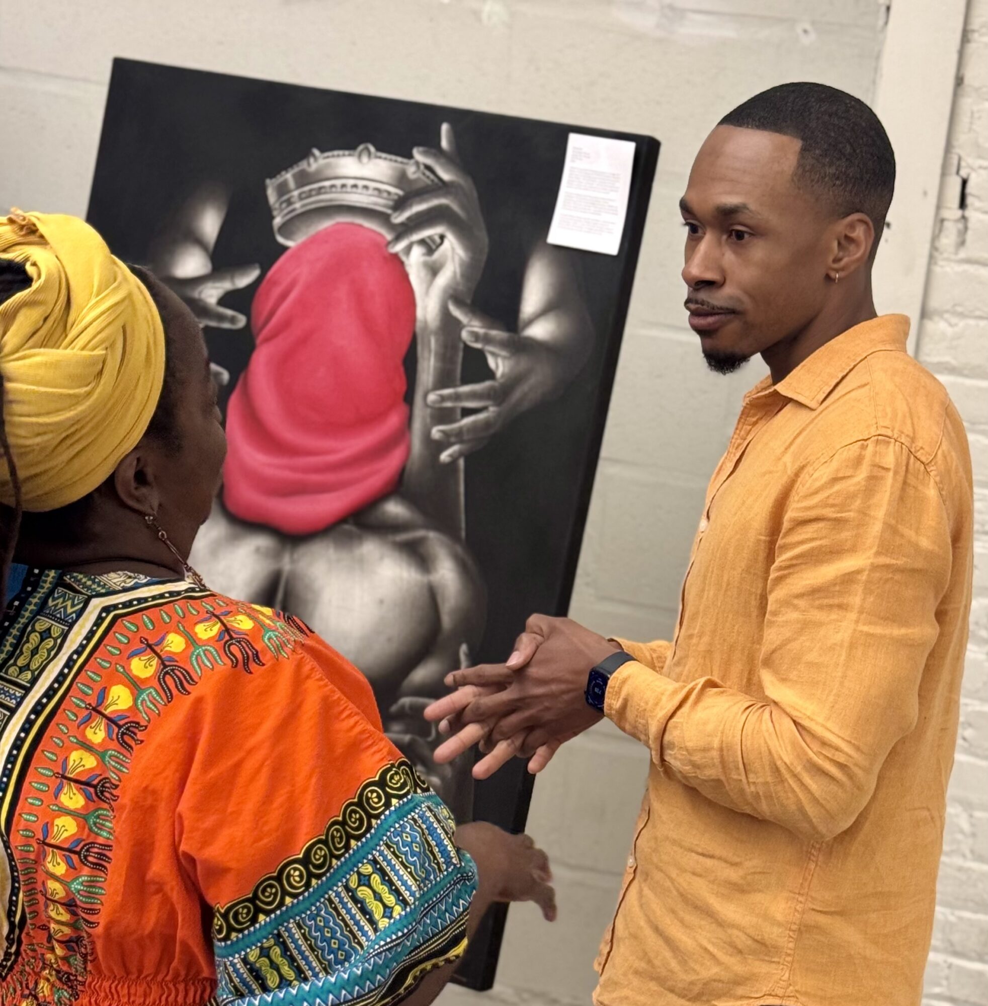 Two people talking in front of a black and white artwork featuring hands and a red cloth, in an indoor setting.
