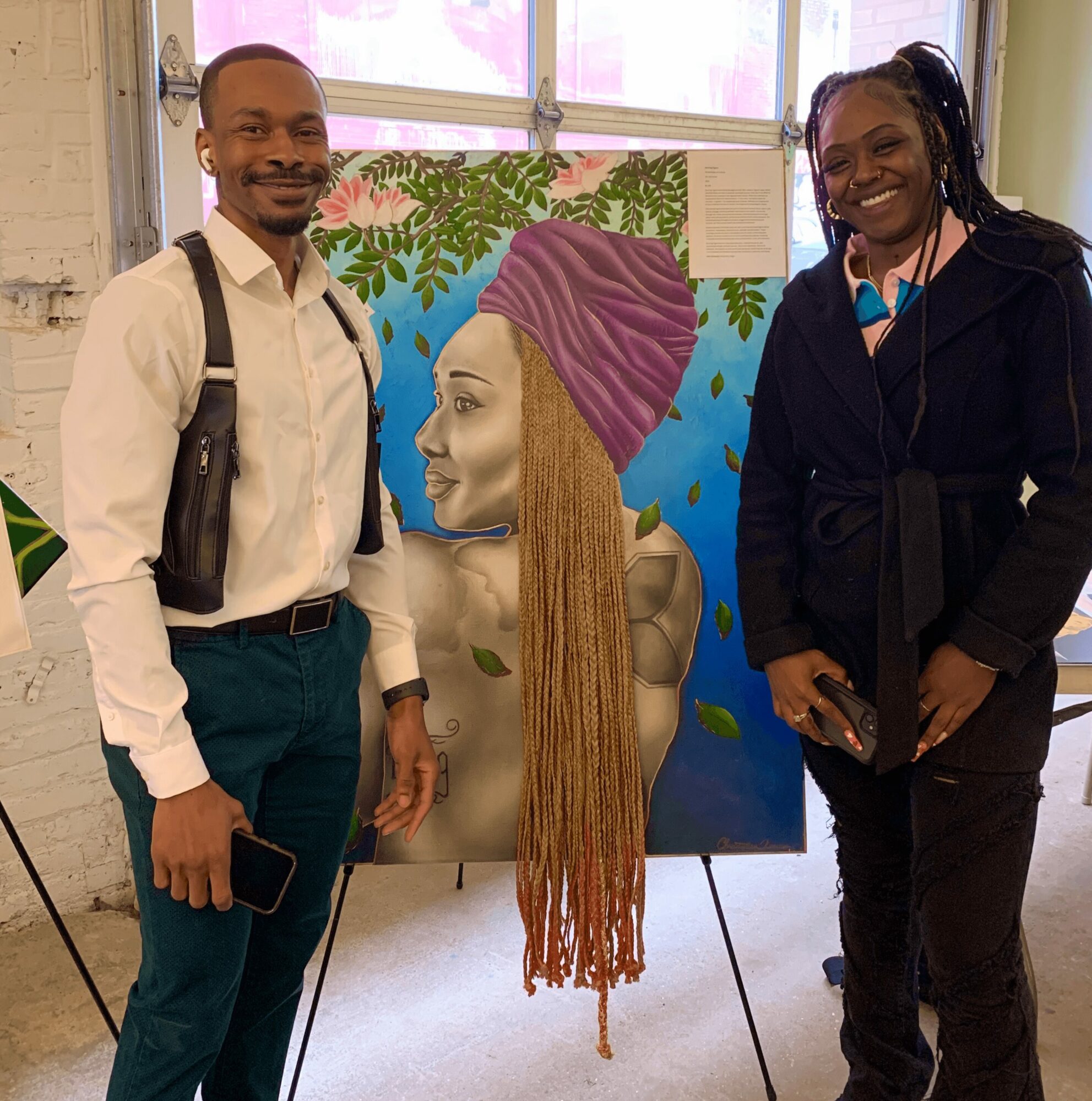Two people stand beside a colorful painting of a woman with long braided hair and a purple headwrap, indoors with large windows.