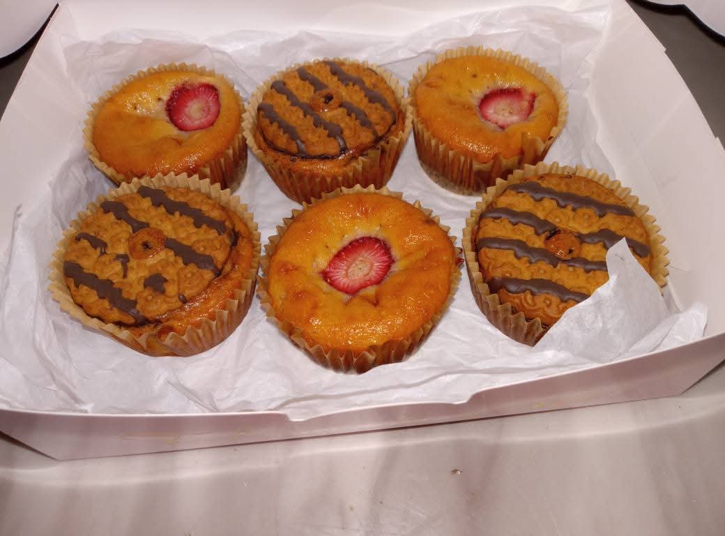 Six cupcakes in a box, some with strawberry toppings and chocolate drizzle, on white paper lining.