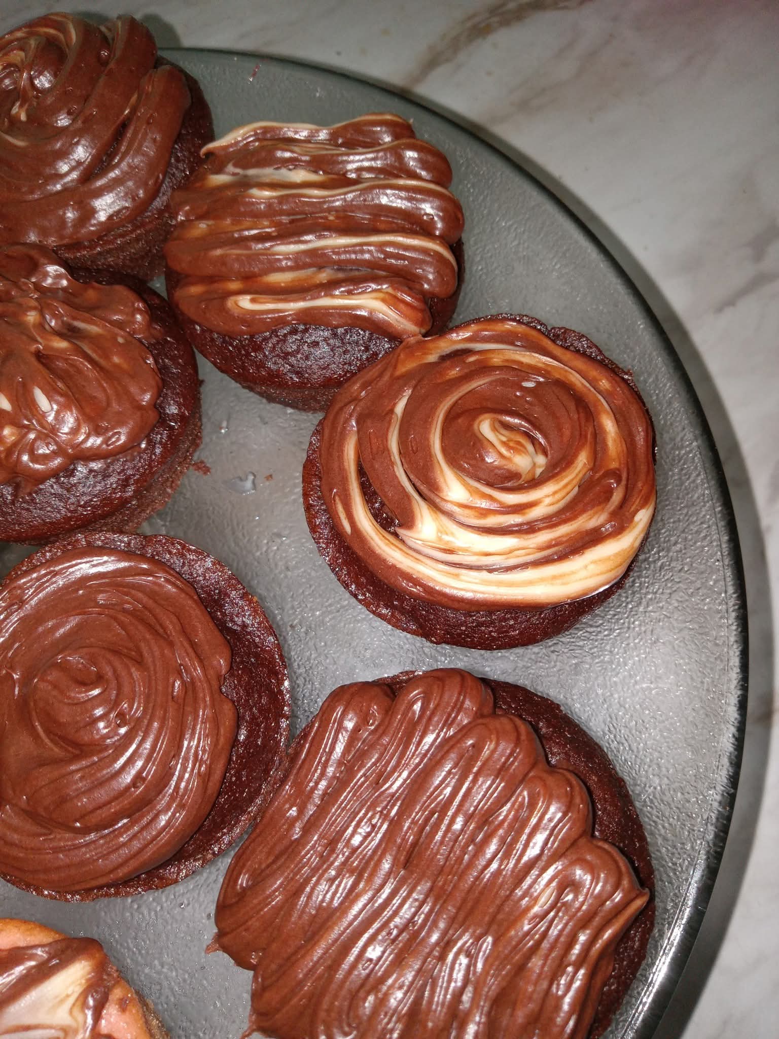 Six chocolate pastries with swirled and textured icing on a gray plate, on a light surface.