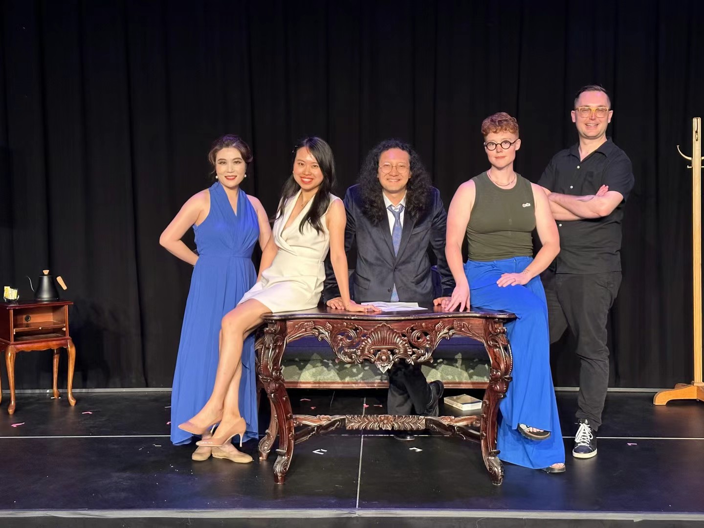 Six people standing on stage behind a decorative table, with a black curtain background, smiling and posing for a photo.