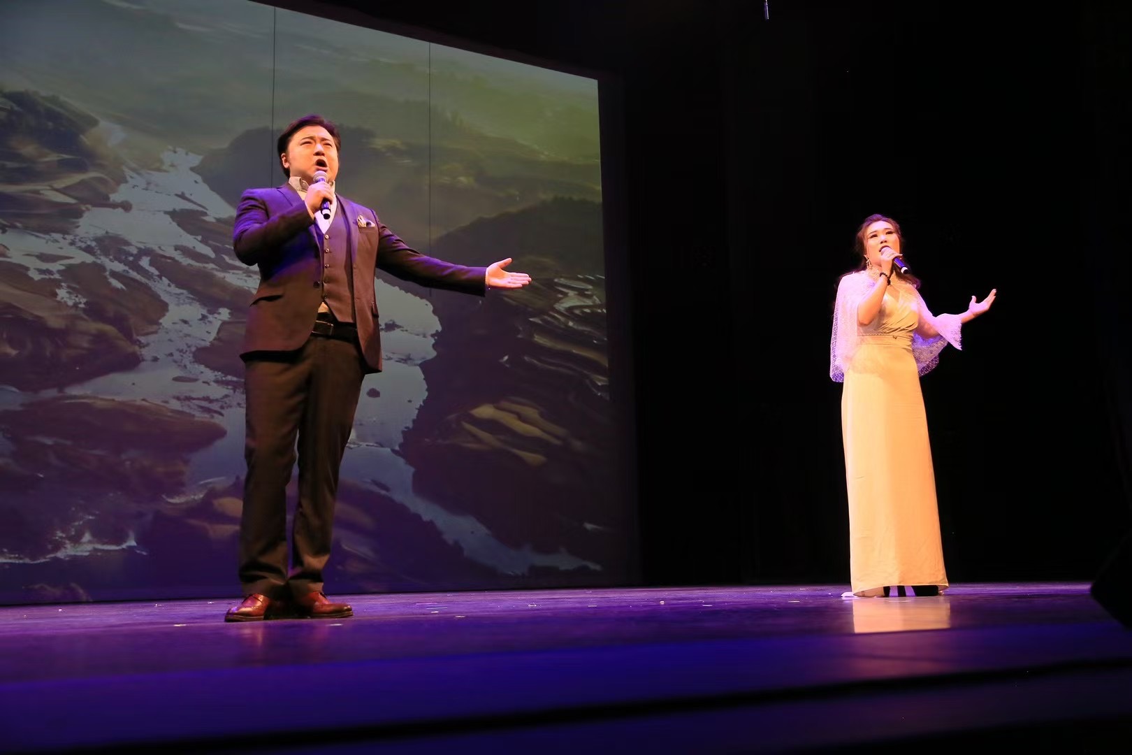 Two performers singing on stage with a scenic background, one in a suit and the other in a long dress.