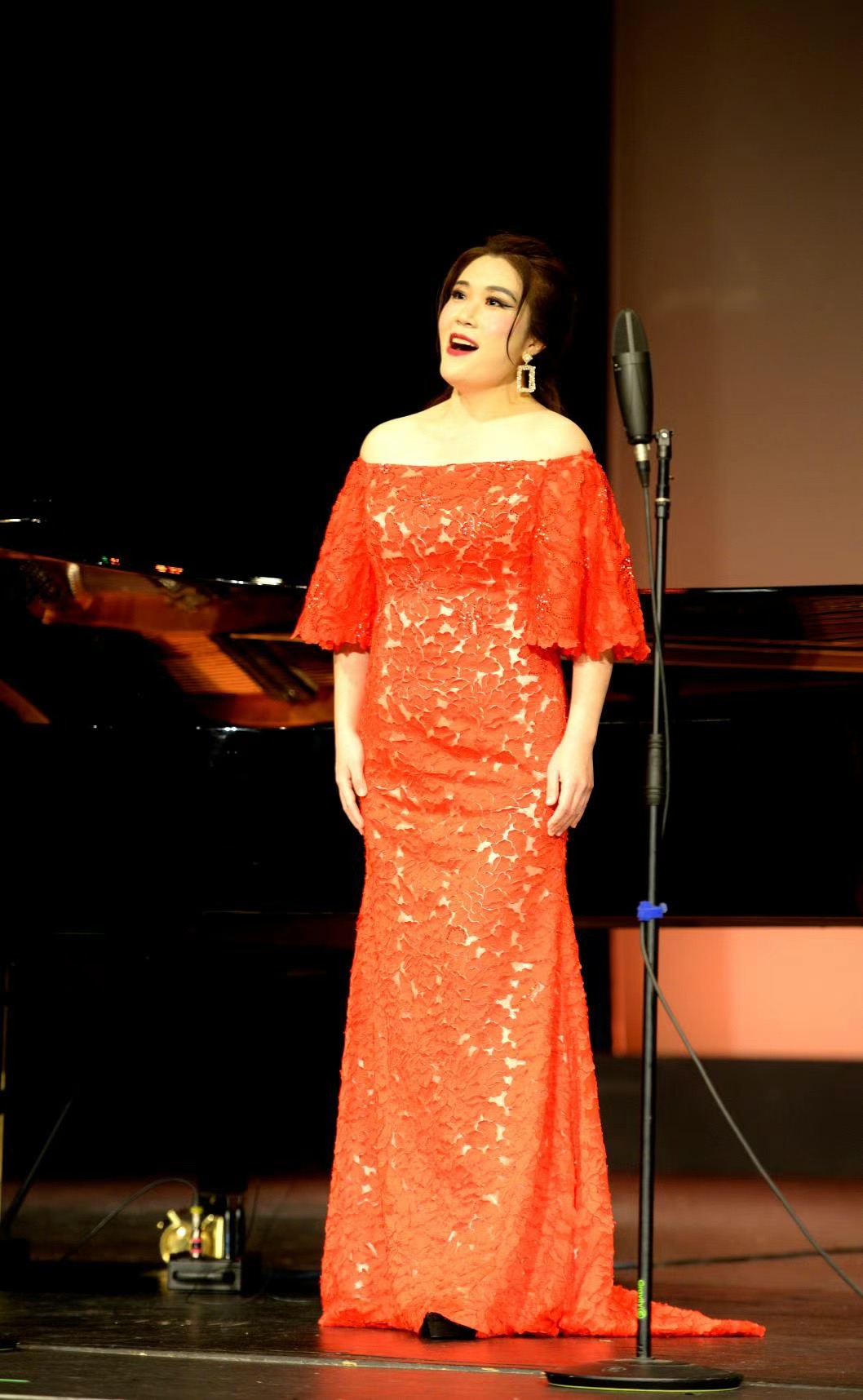 Woman in red gown singing on stage with microphone, piano in background, stage lighting, smiling, elegant attire.