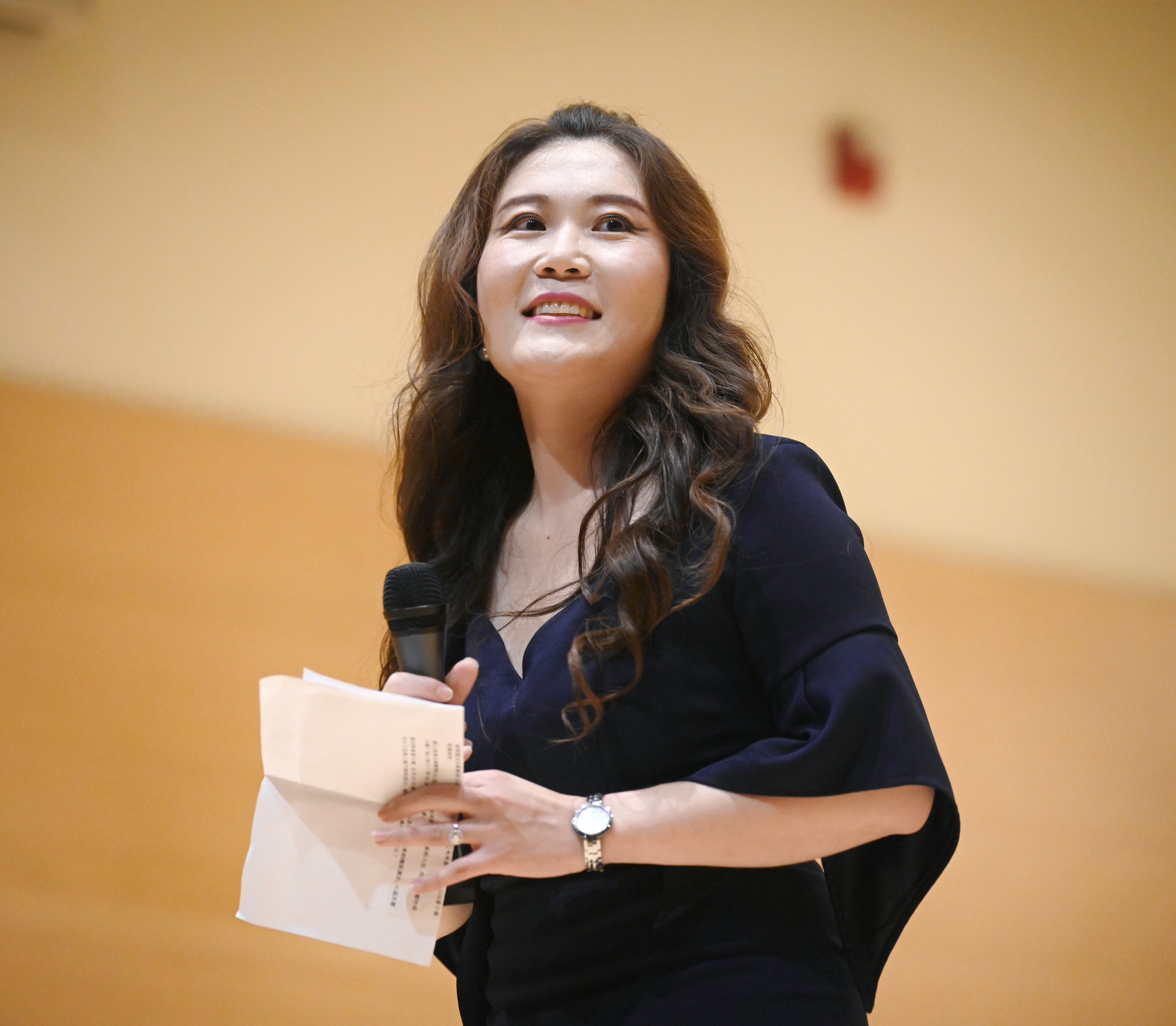 Woman with long wavy hair holding papers and a microphone, smiling, in an indoor setting.
