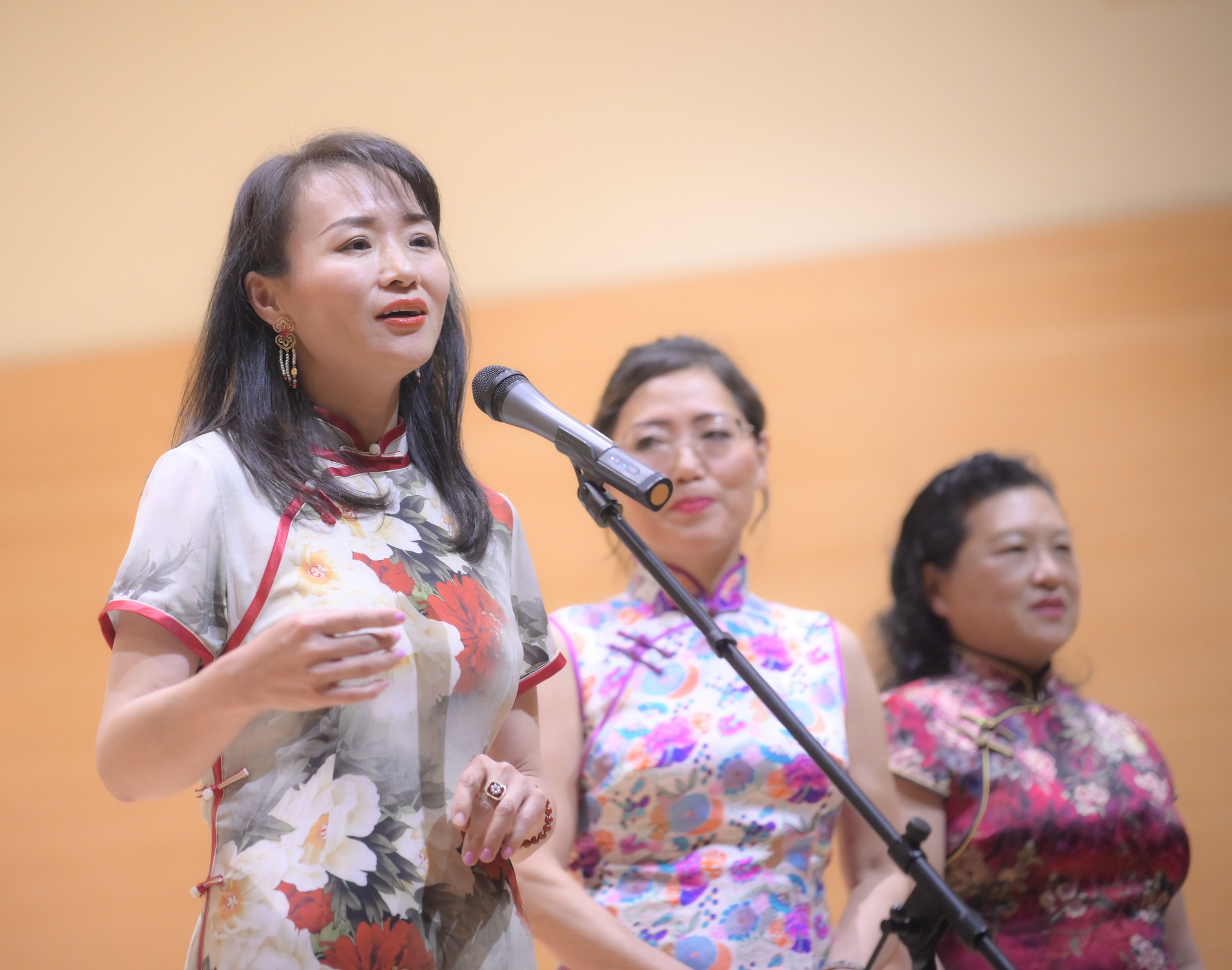 Three women standing on stage, one speaking into a microphone, with a plain background.