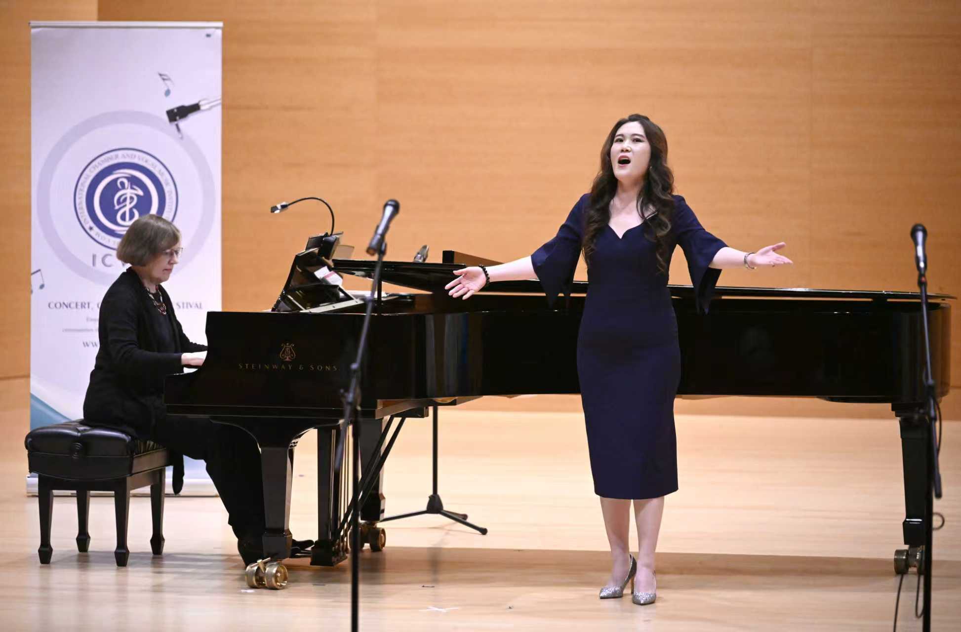 A woman sings with arms outstretched while a pianist plays on a grand piano in a concert hall.