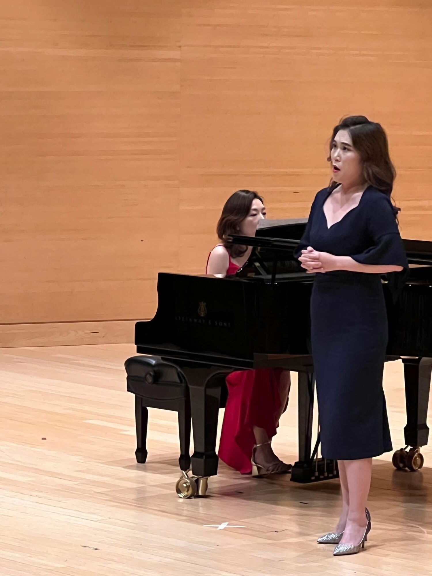 Woman singing with a pianist in a concert hall, both dressed formally, wooden wall background.