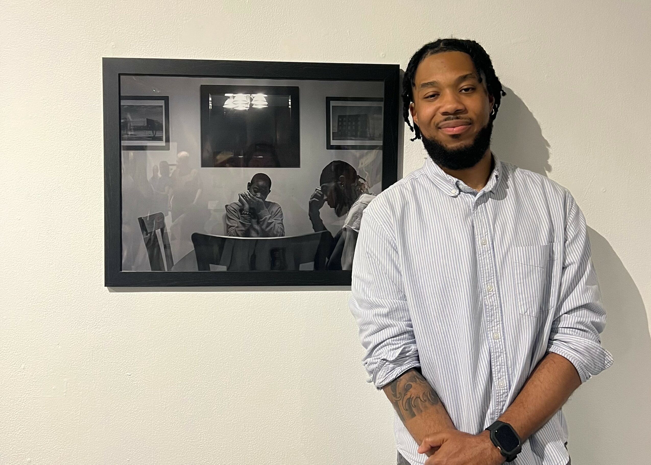 Young man with a beard and tattoos, wearing a light-colored shirt, standing in front of a wall with a framed mirror reflecting two people, one with a striped shirt, in a room with chairs.