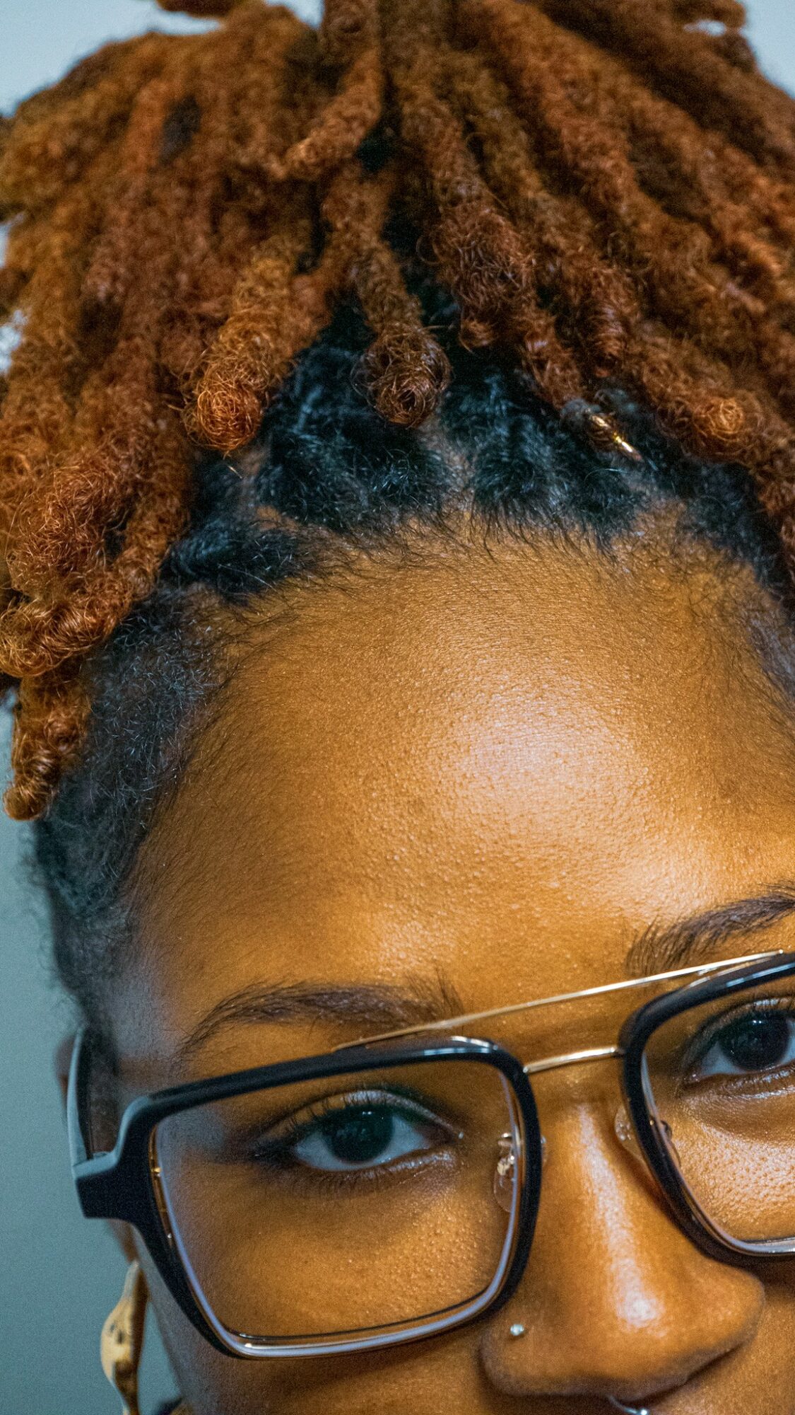 Close-up of a person with brown dreadlocks, wearing glasses and earrings, with a neutral background.