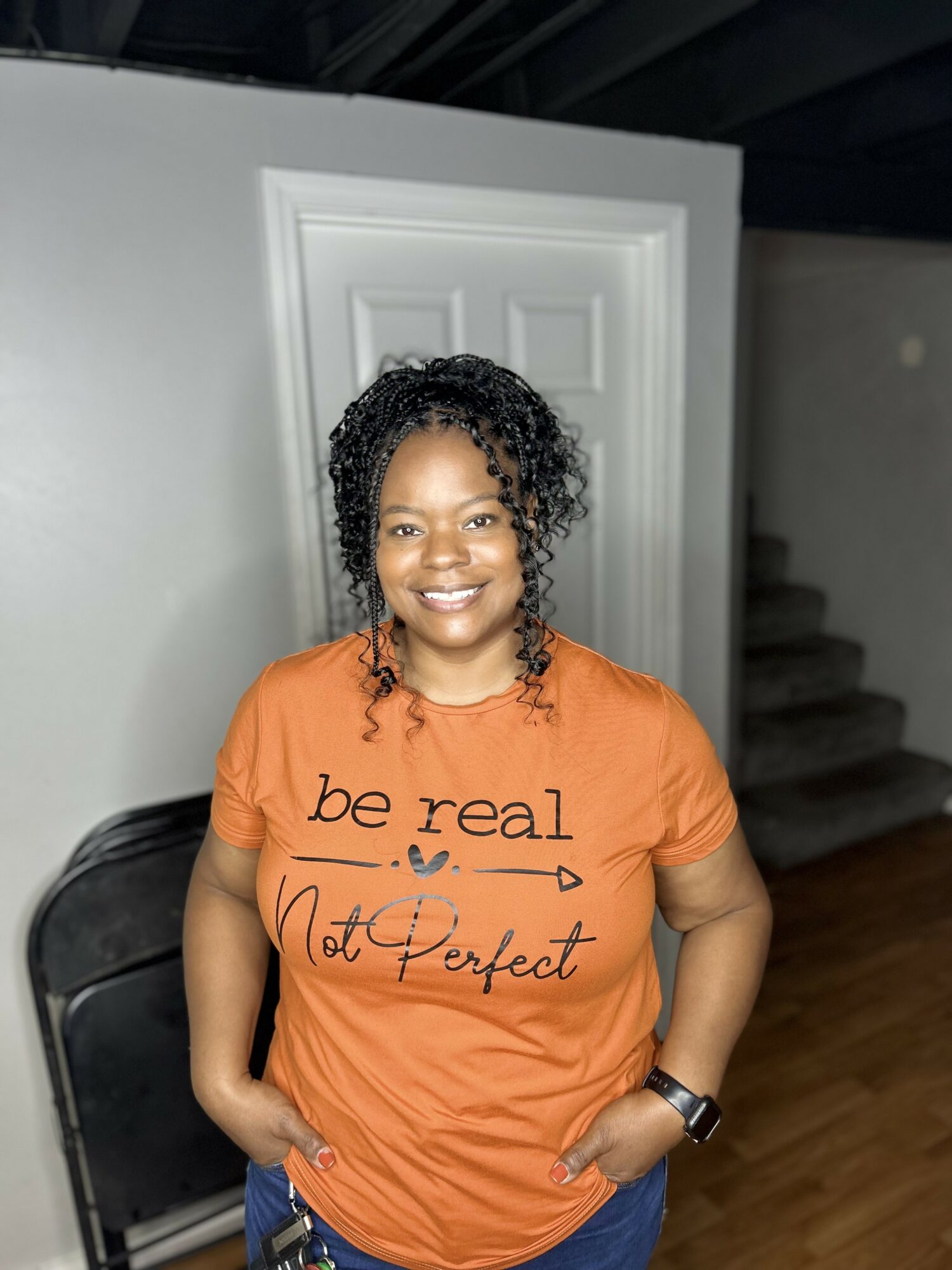 Smiling woman with curly hair wearing an orange t-shirt with text, standing indoors near a door and staircase.
