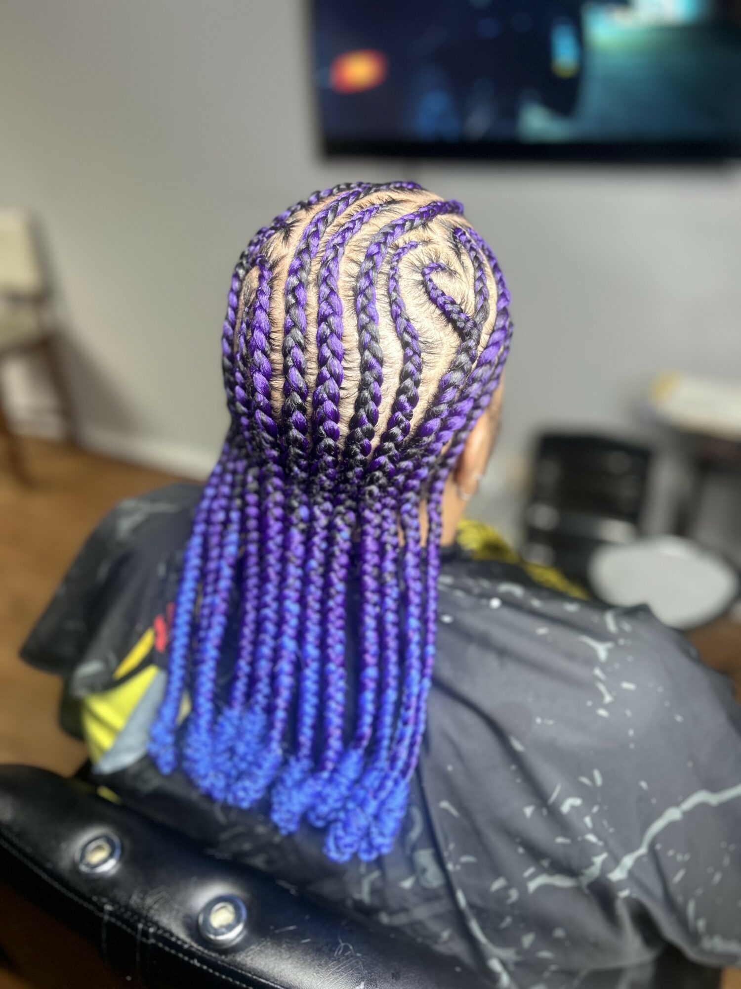 Person with colorful braided hair, transitioning from purple to blue, sitting in a salon chair, facing away from the camera.