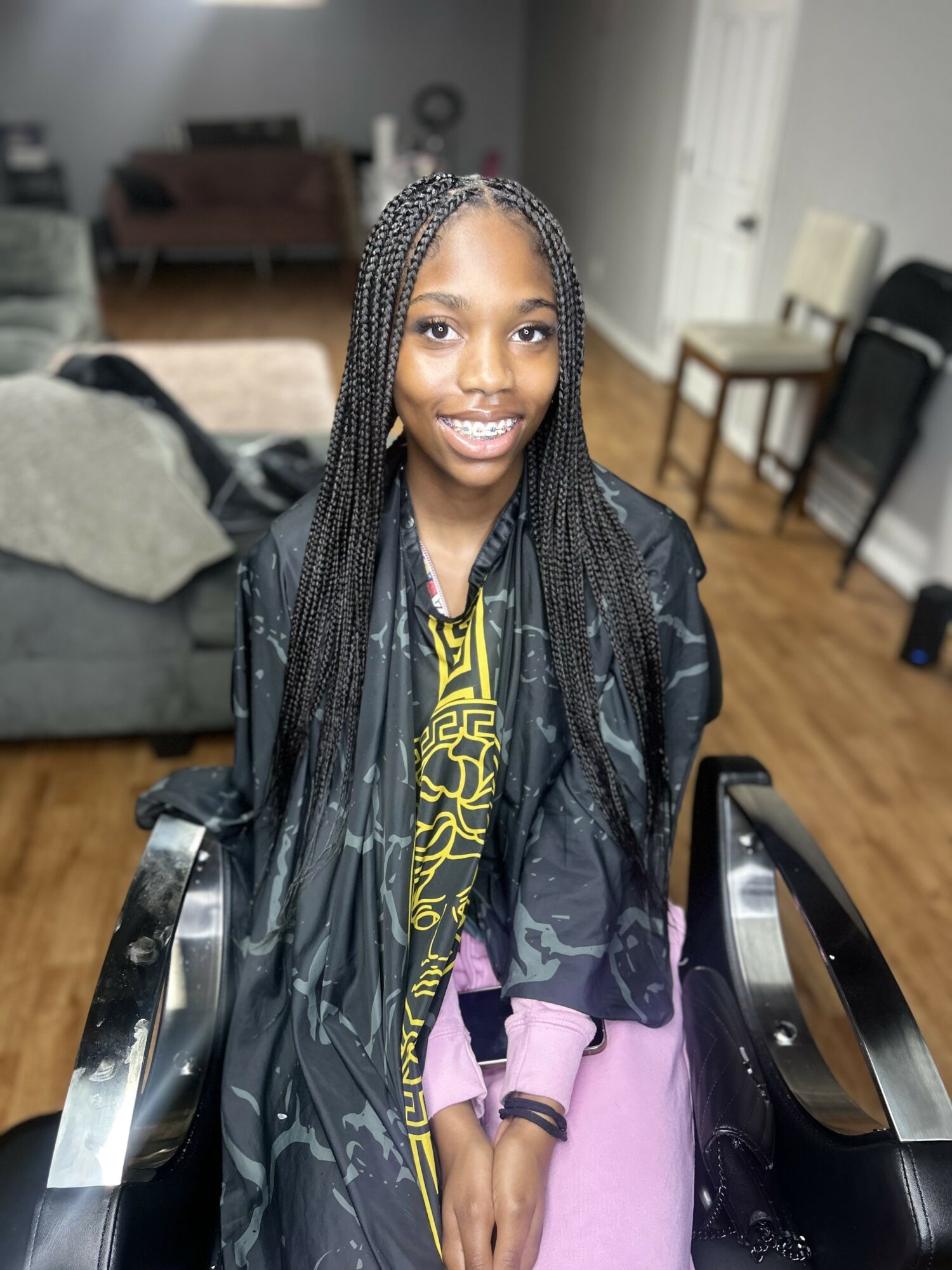 Young girl with braided hair smiling, standing in a living room with furniture and hardwood floor.