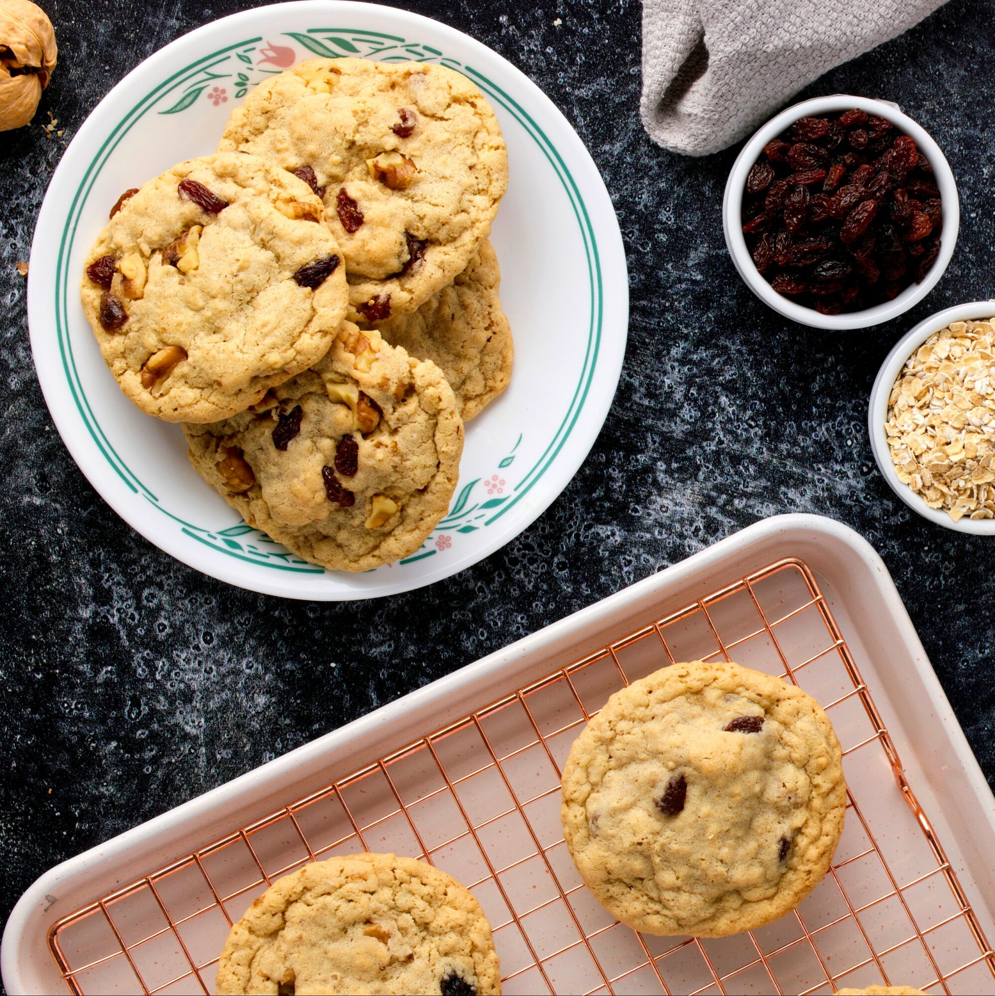 Three chocolate chip cookies on a white plate, with additional cookies on a cooling rack, on a dark countertop.