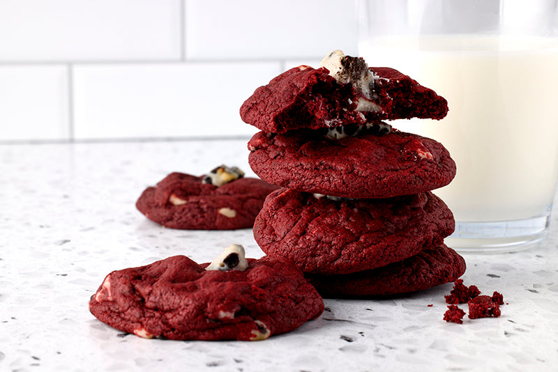 Stack of four red cookies with chocolate chips on a white surface, with a glass of milk in the background.