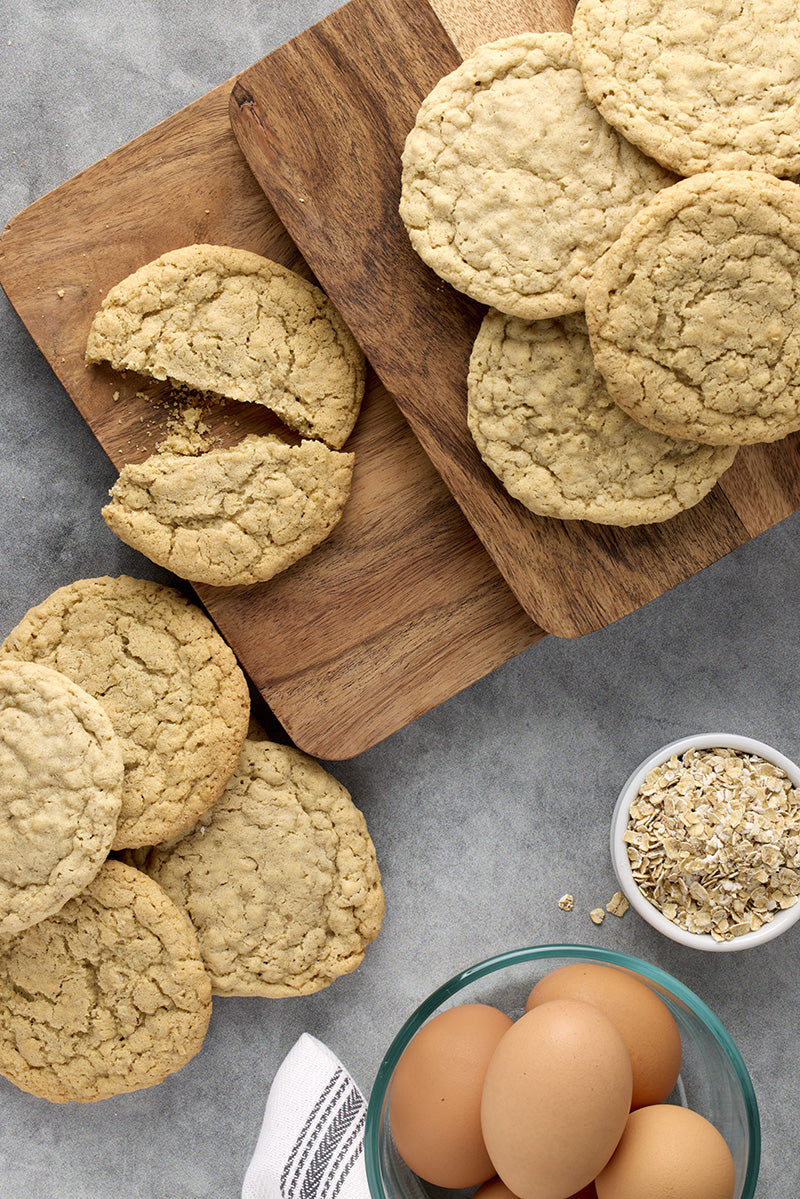 Several oatmeal cookies on a wooden cutting board and a few on a gray surface, with a bowl of oats and eggs nearby.