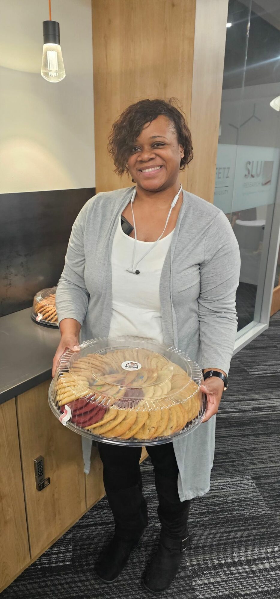 Woman smiling holding a large tray of assorted cookies in a modern indoor setting.