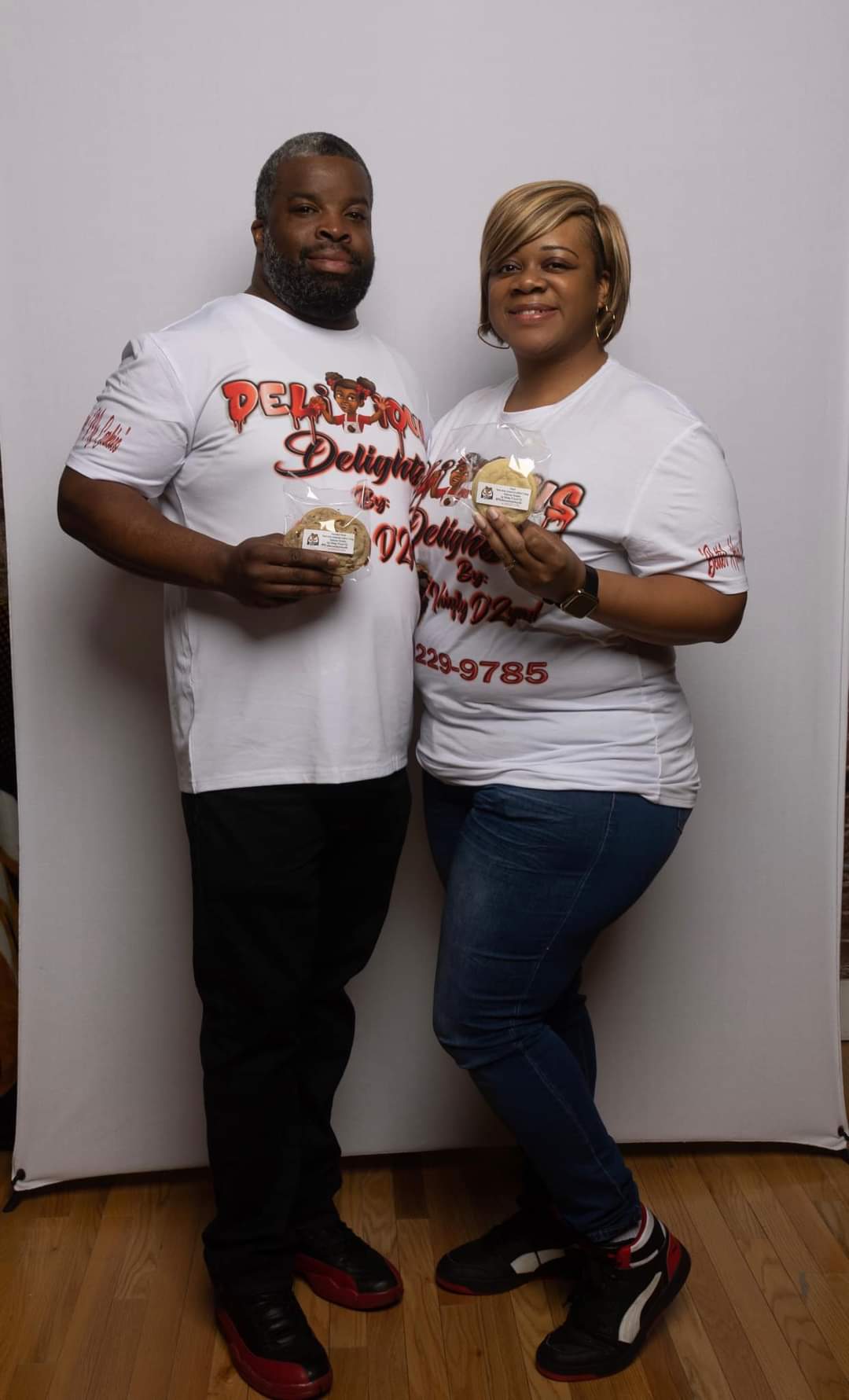 Two people stand side by side holding food items, smiling, against a plain white background.