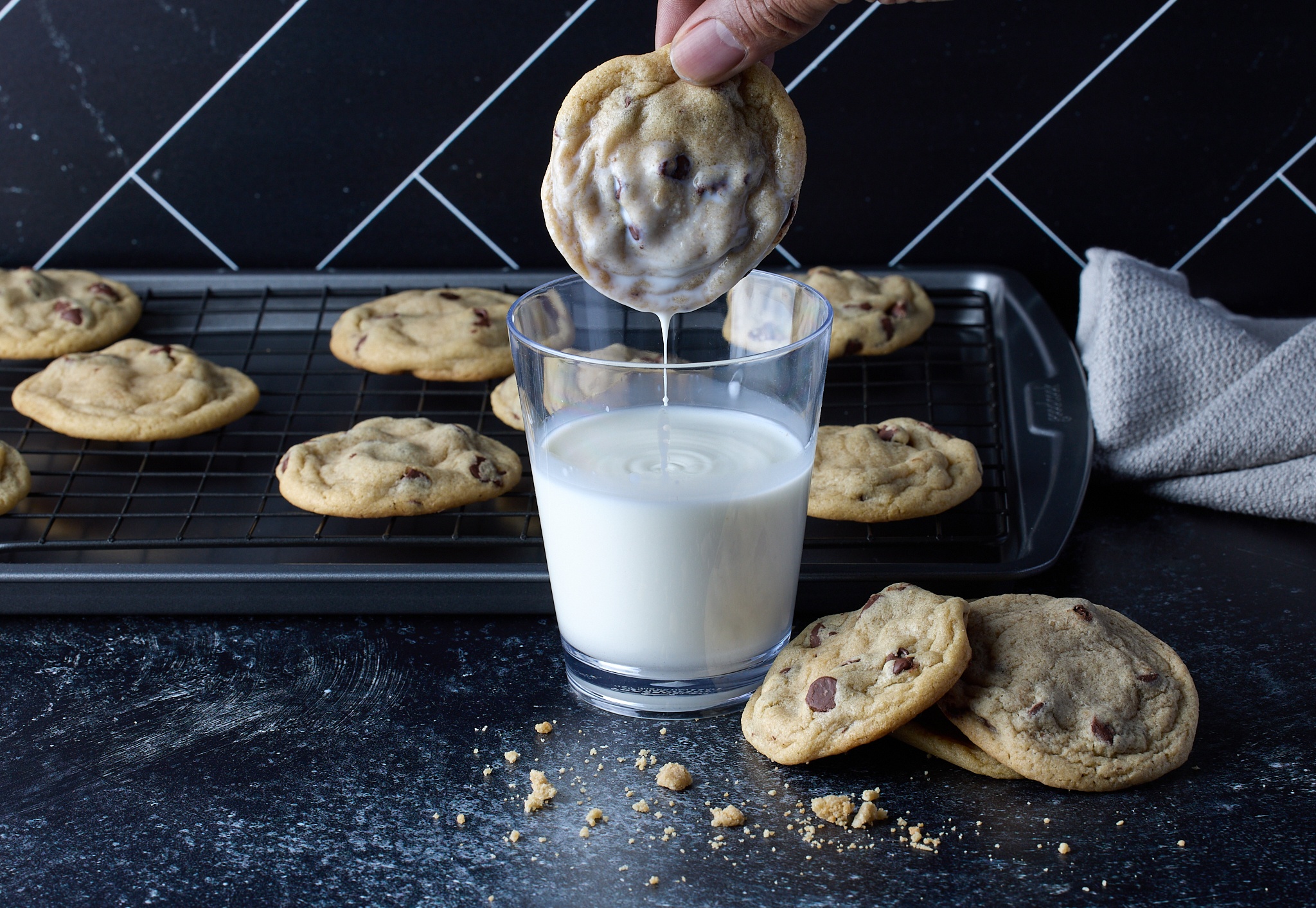 Hand dips chocolate chip cookie into a glass of milk, with cookies on a cooling rack and scattered crumbs nearby.