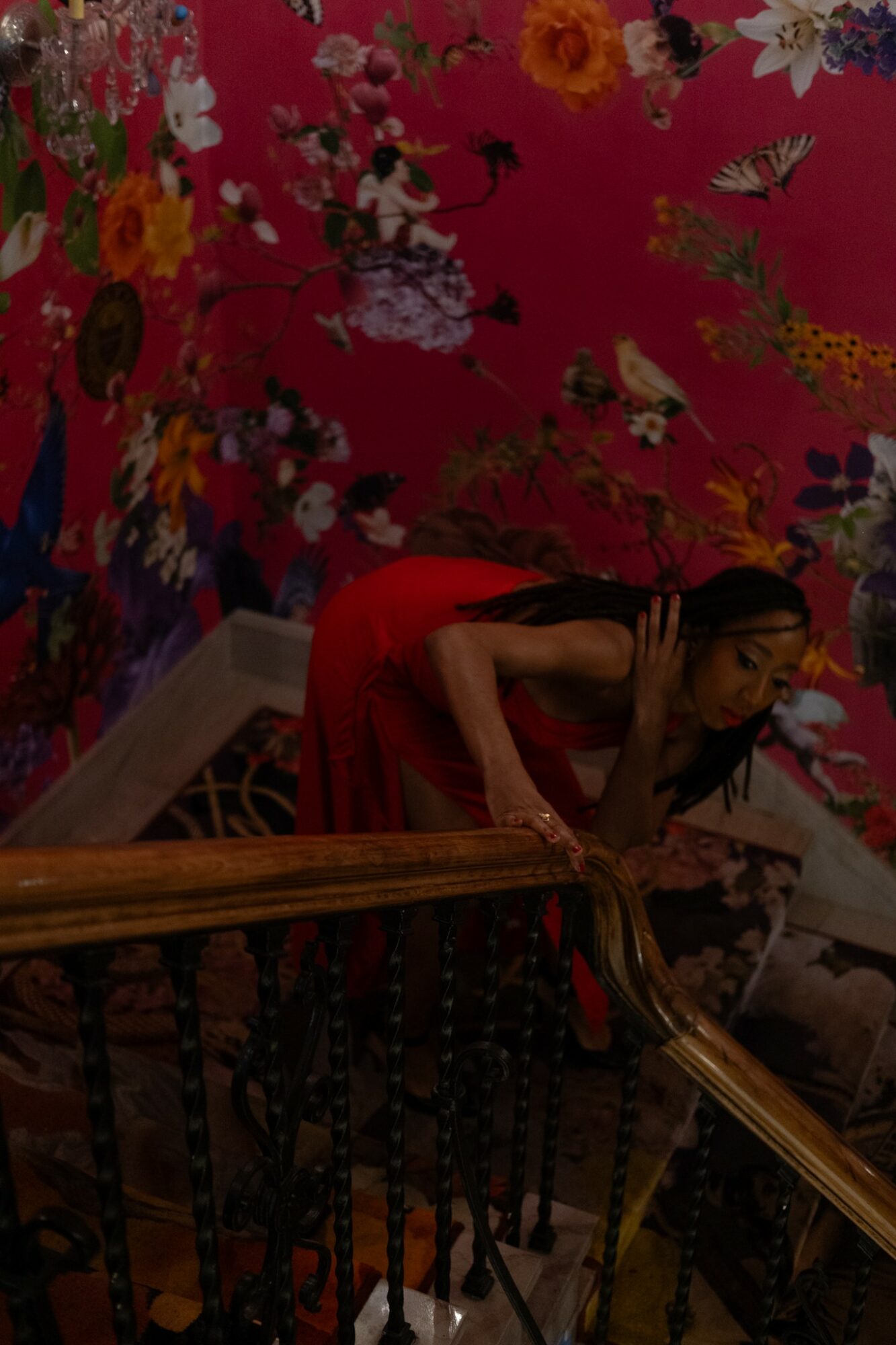 Woman in red dress descending staircase with floral wallpaper background.