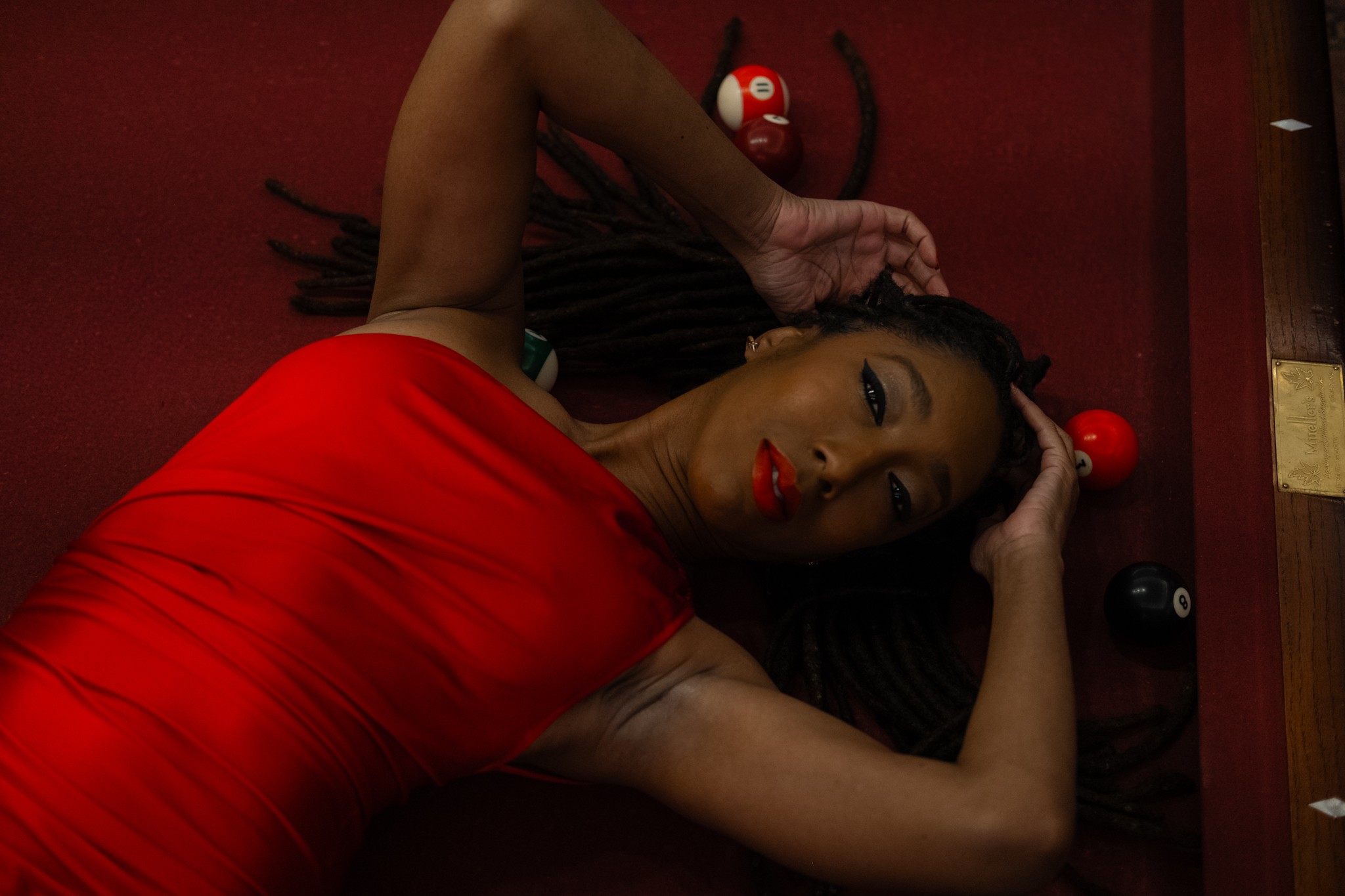 Woman lying on red surface, wearing red dress, with black hair and makeup, surrounded by small red and white objects.