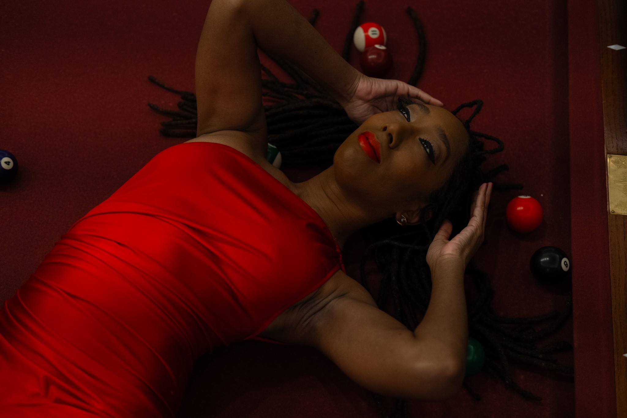 Woman lying on a red surface surrounded by poker chips, wearing a red dress, with her eyes closed and hands near her head.