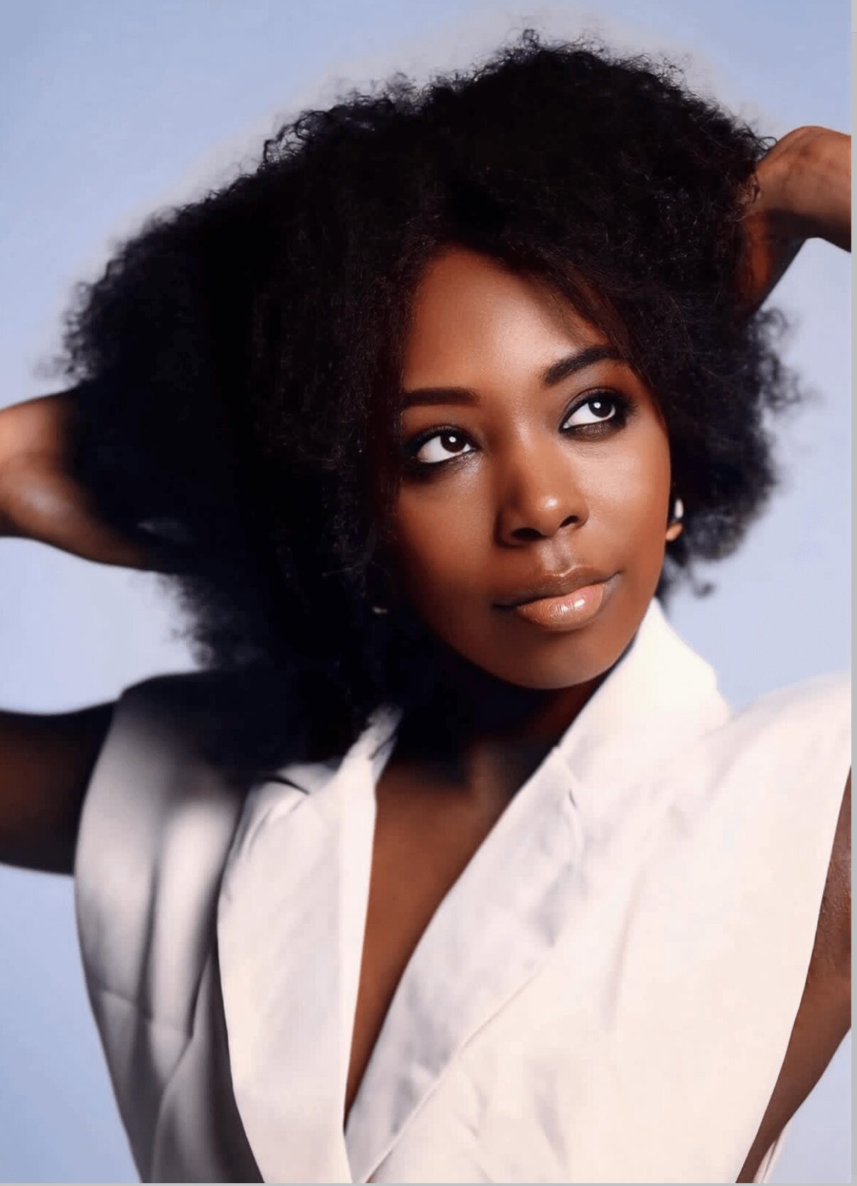 Woman with natural curly hair looking to the side, wearing a white top, with hands behind her head.