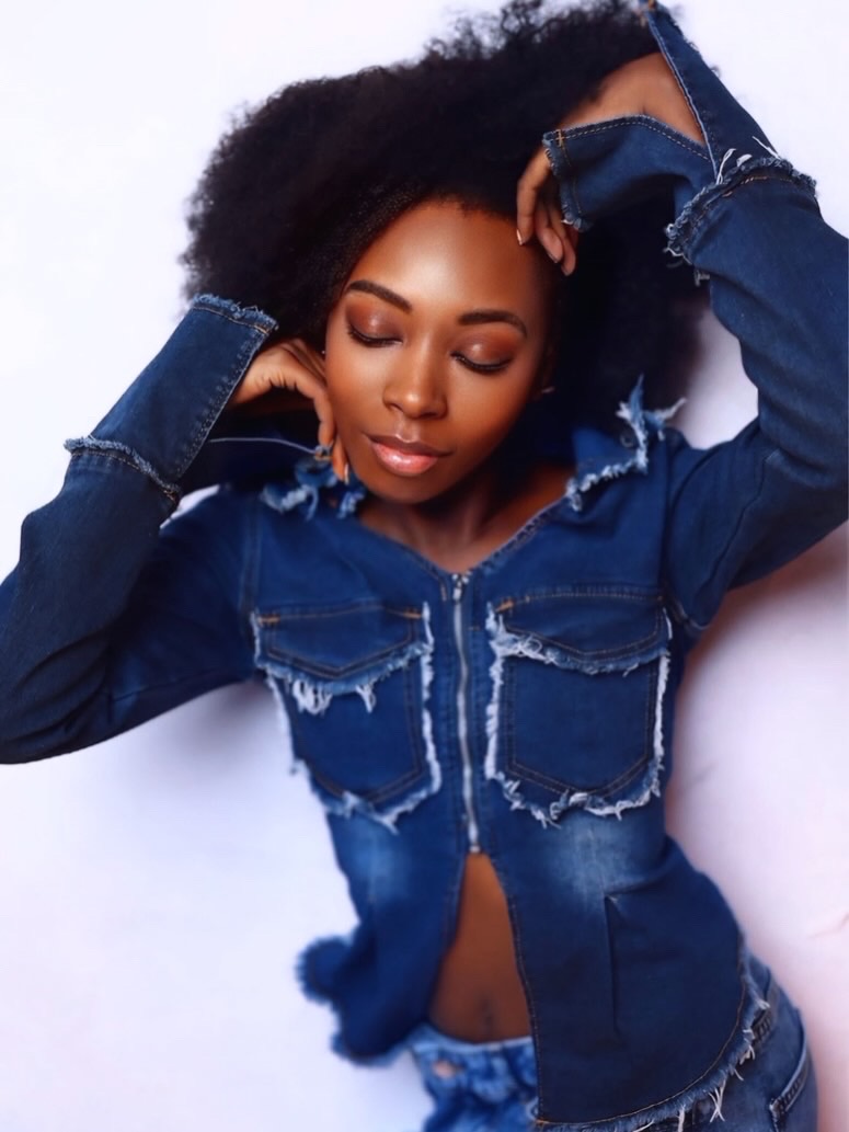 Portrait of a woman with natural hair, wearing a ripped denim jacket, eyes closed, resting her head on her hands.