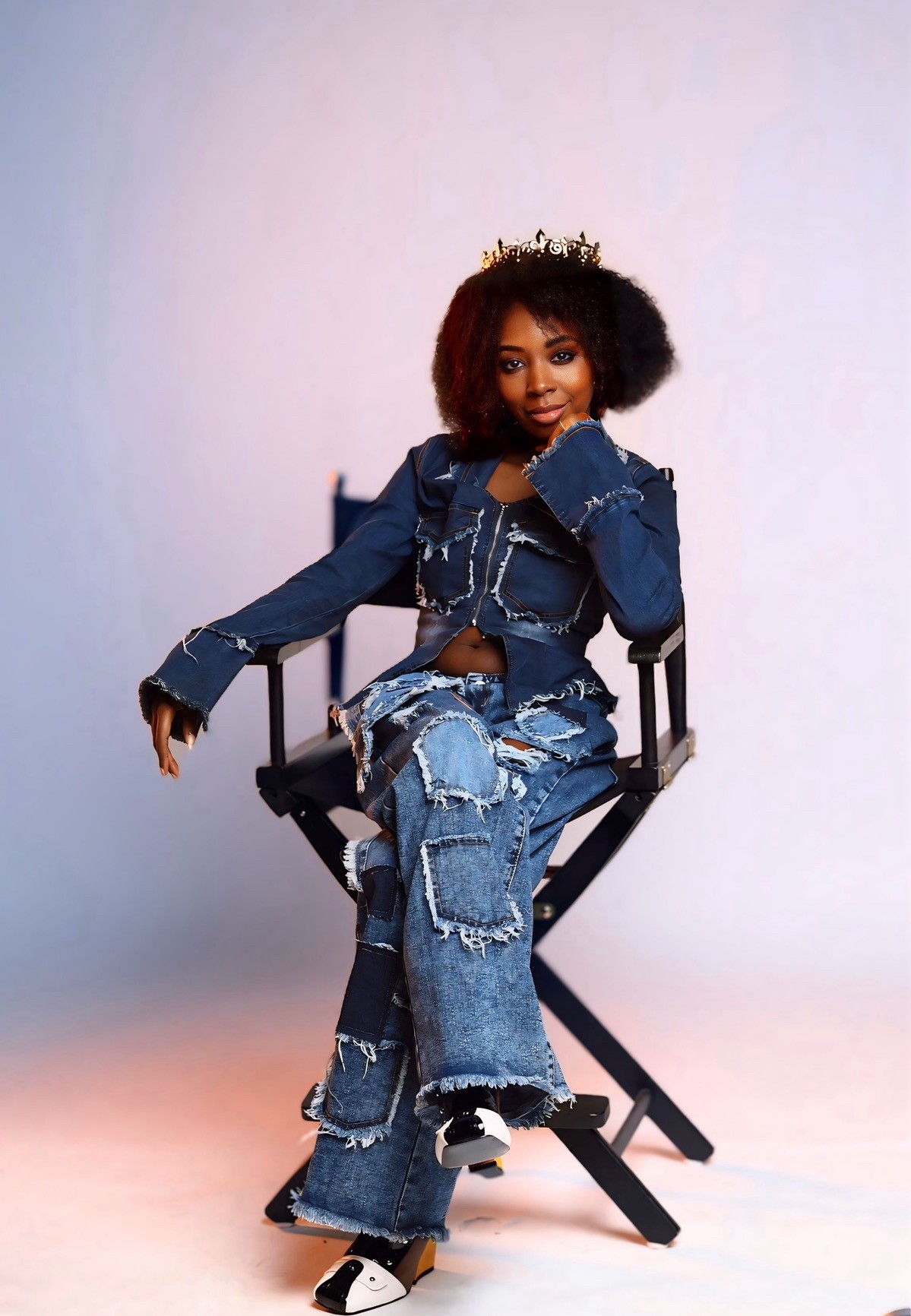 Young woman with curly hair wearing a denim outfit and a crown, sitting on a director's chair against a plain background.